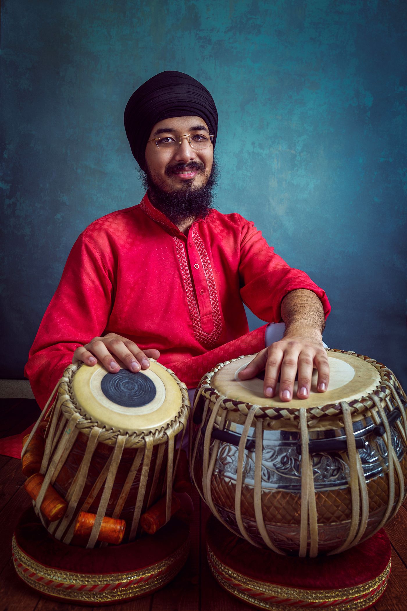 Man in a red traditional outfit and turban sitting with tabla and dholak, smiling against a textured blue background.