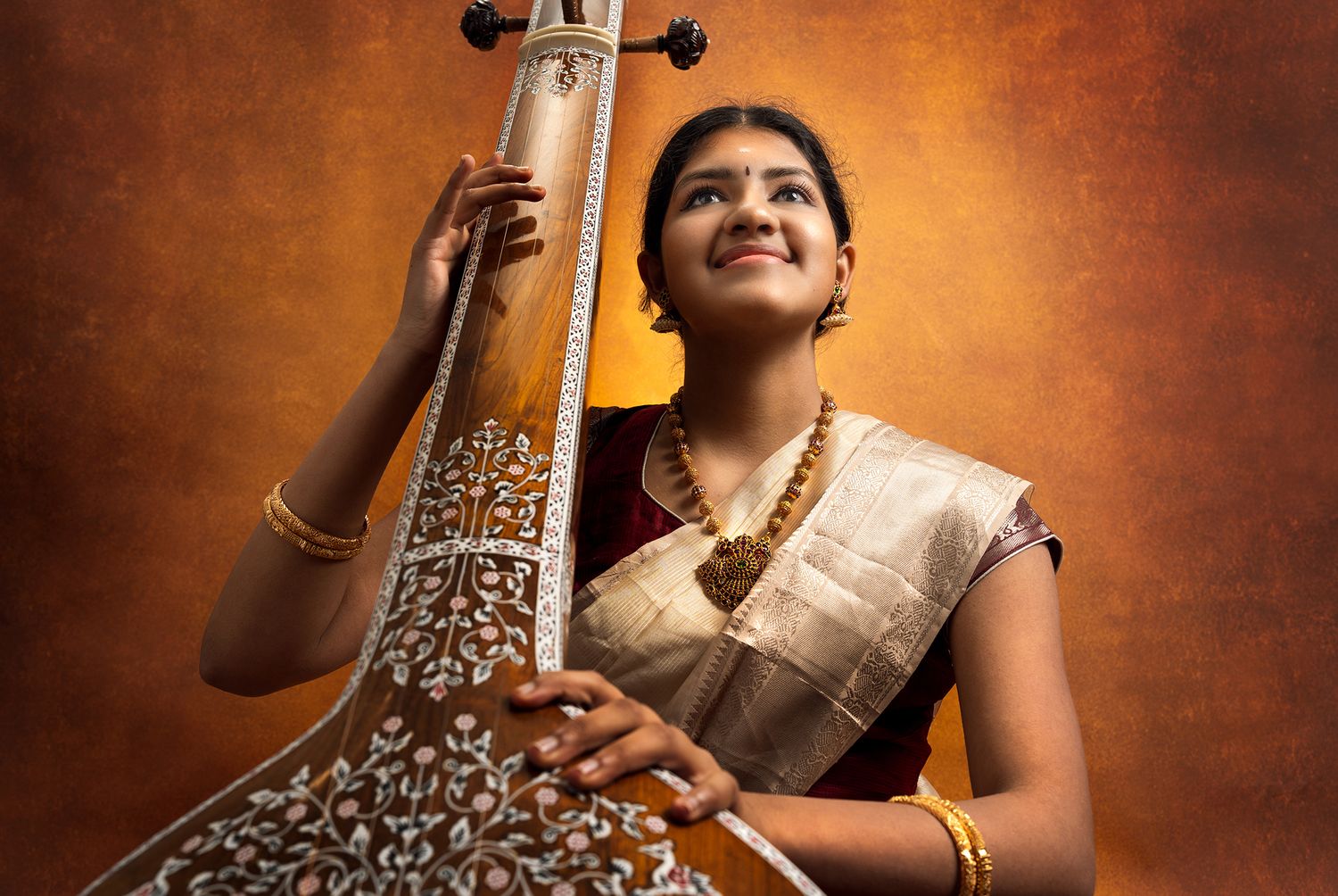Young woman in traditional attire holding a tanpura, wearing gold jewelry, against a warm, textured background.