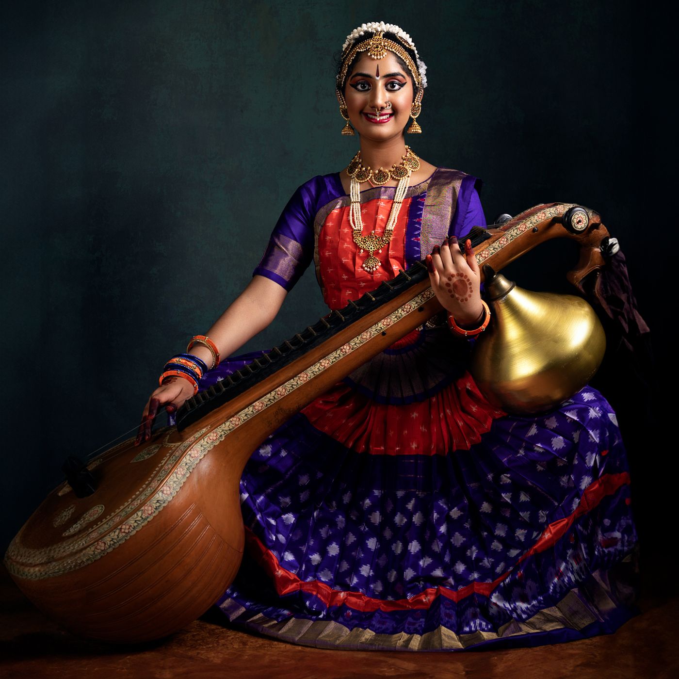A woman dressed in traditional attire playing a musical instrument, likely a sarangi. She is seated, wearing a colorful outfit with intricat