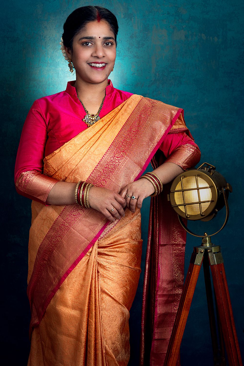 Woman wearing elegant orange silk saree poses with vintage globe against teal background.