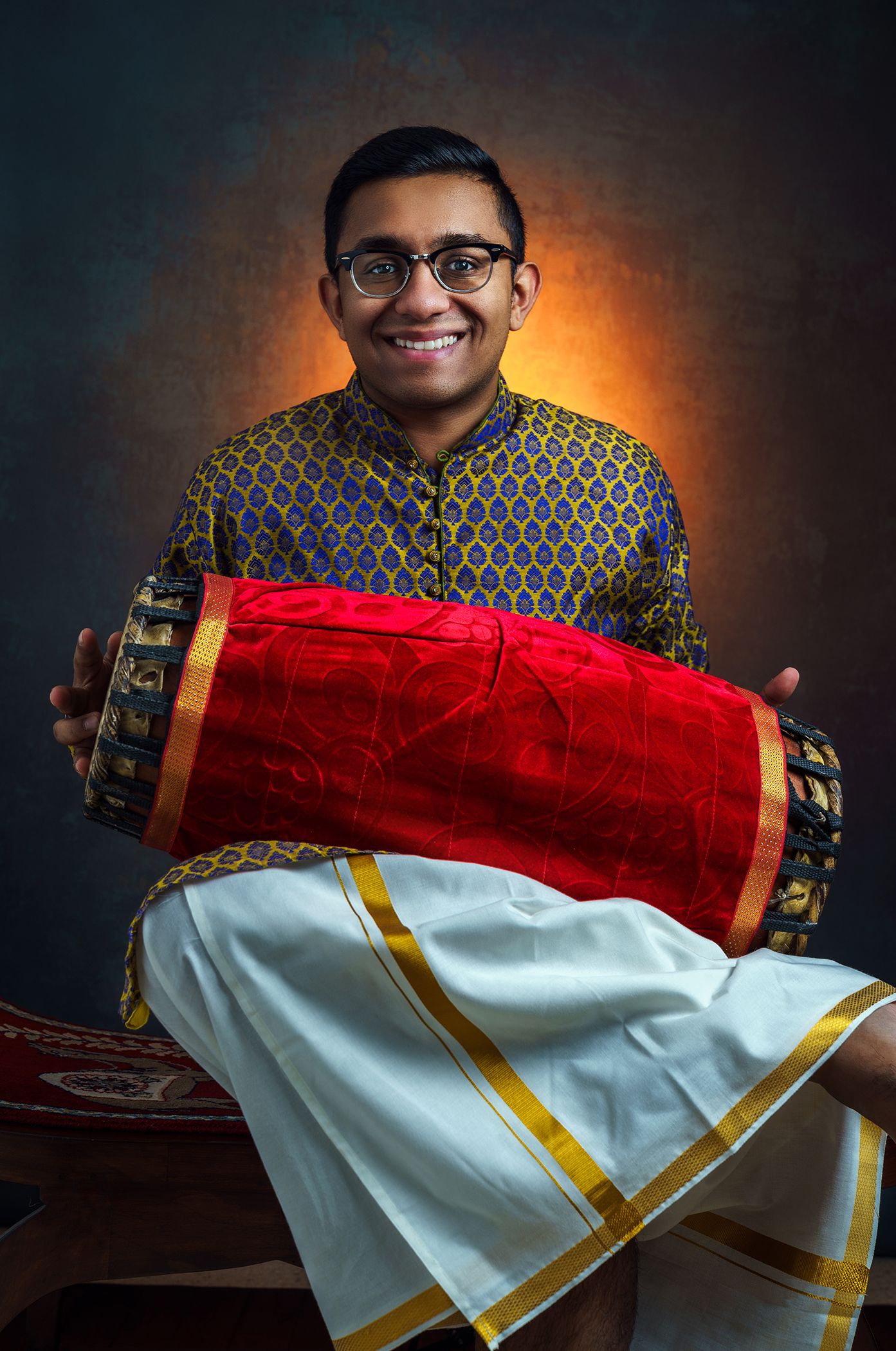 Young man in glasses and a blue patterned kurta holding a red drum, wearing a white dhoti with gold border against a dark, warm-lit backgrou