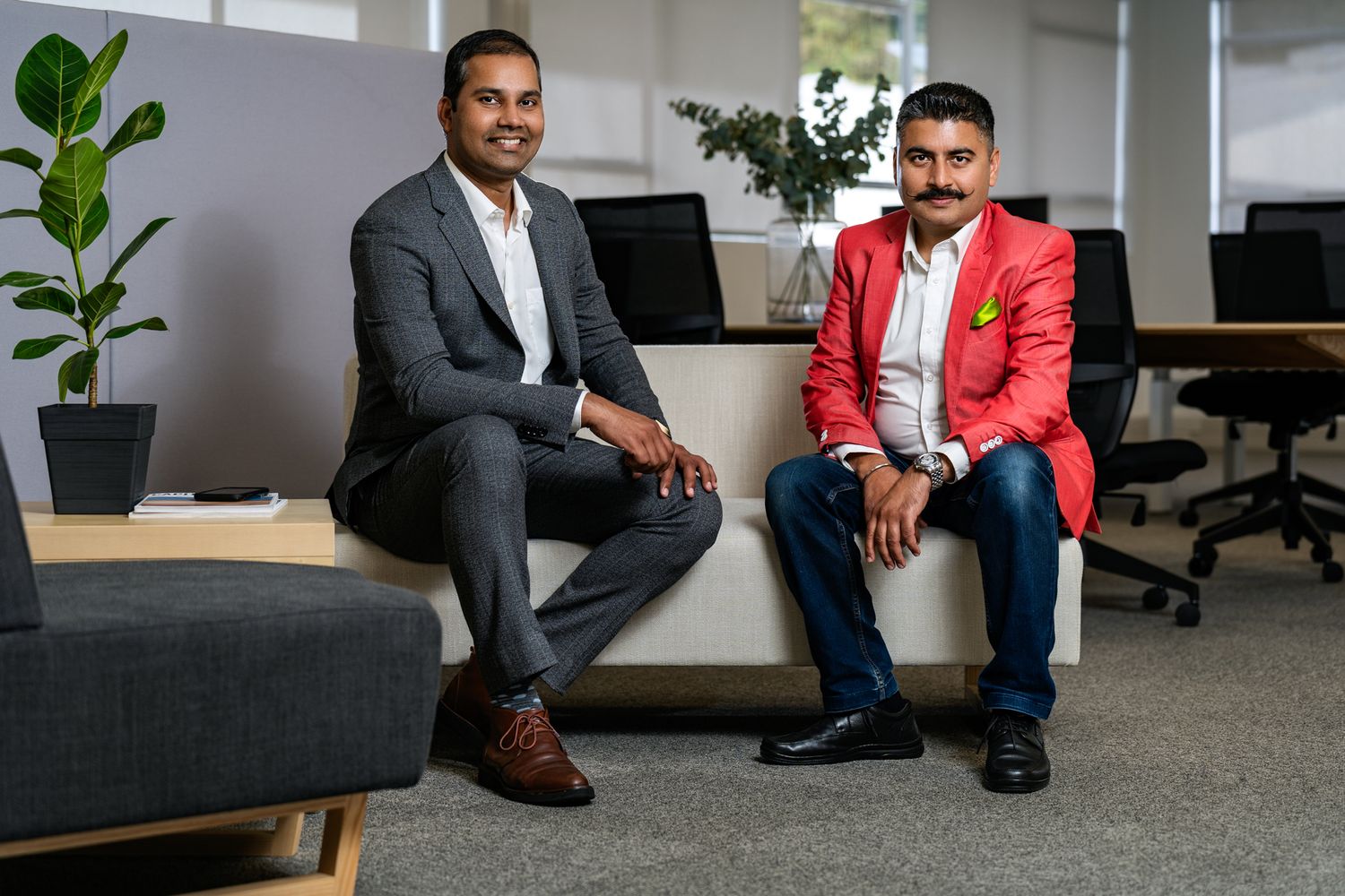 Two men seated in an office, one in a gray suit and the other in a red blazer, with modern furniture and a plant in the background.