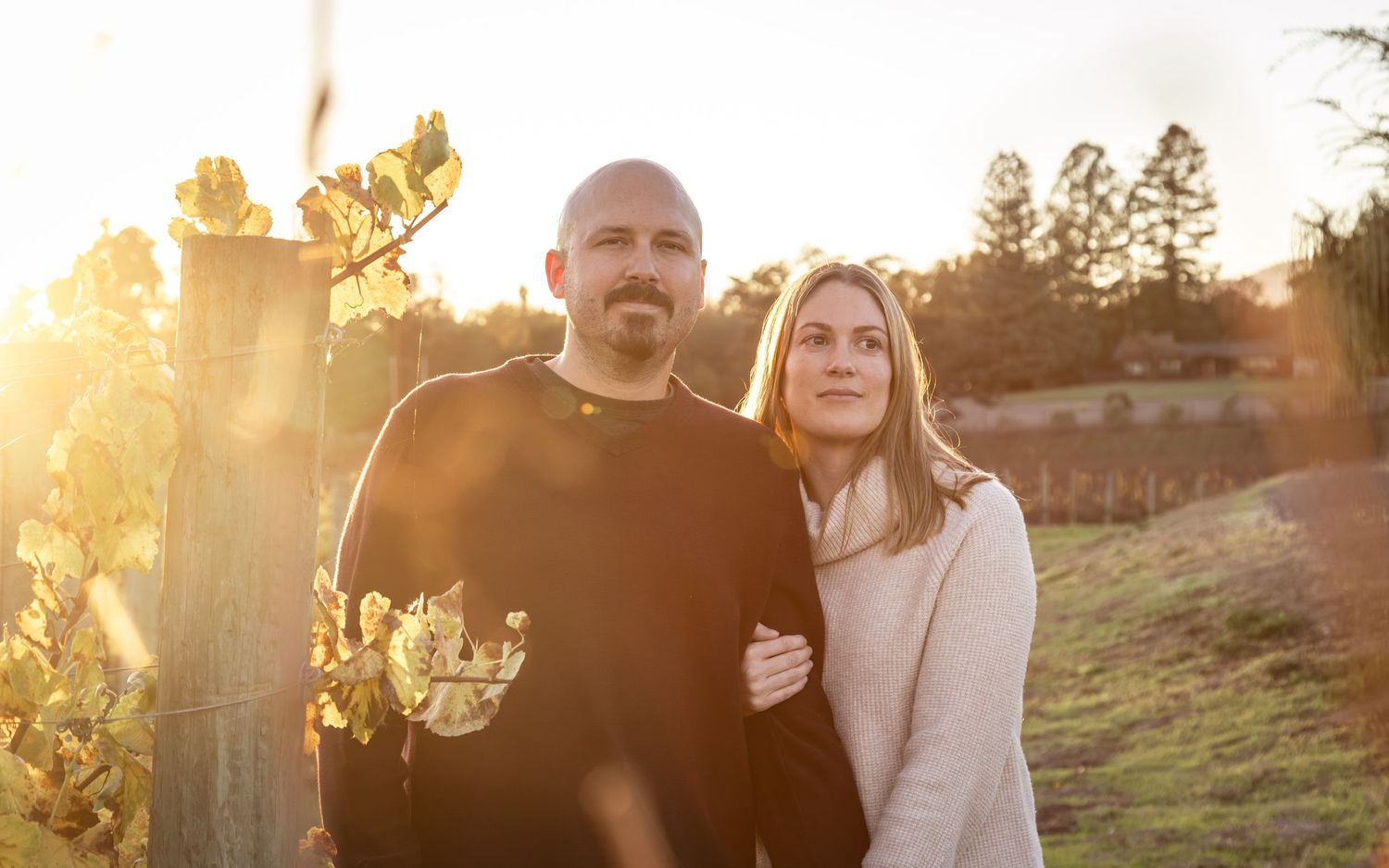 Two people stand together in a vineyard at sunset, bathed in golden light, surrounded by grapevines and a serene landscape.