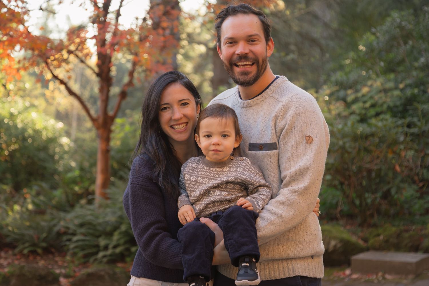 Two adults and a child stand close together in a lush, green outdoor setting, framed by autumn trees and golden light.
