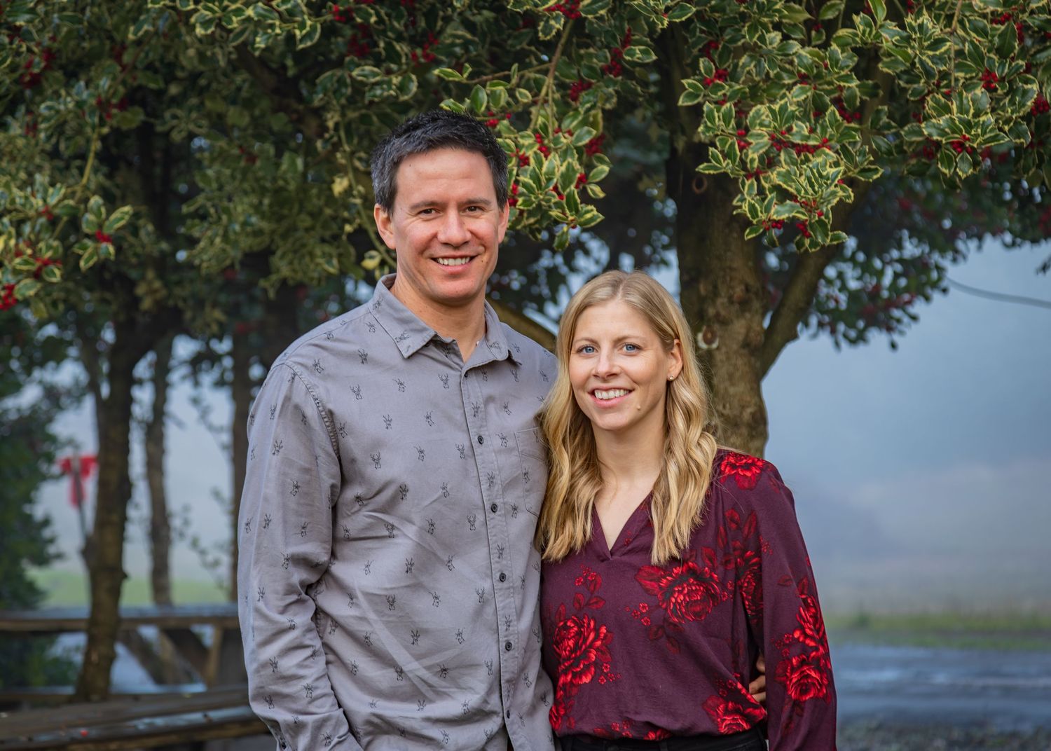 Two people stand together near a tree adorned with red berries, surrounded by misty scenery and a wooden fence in the background.