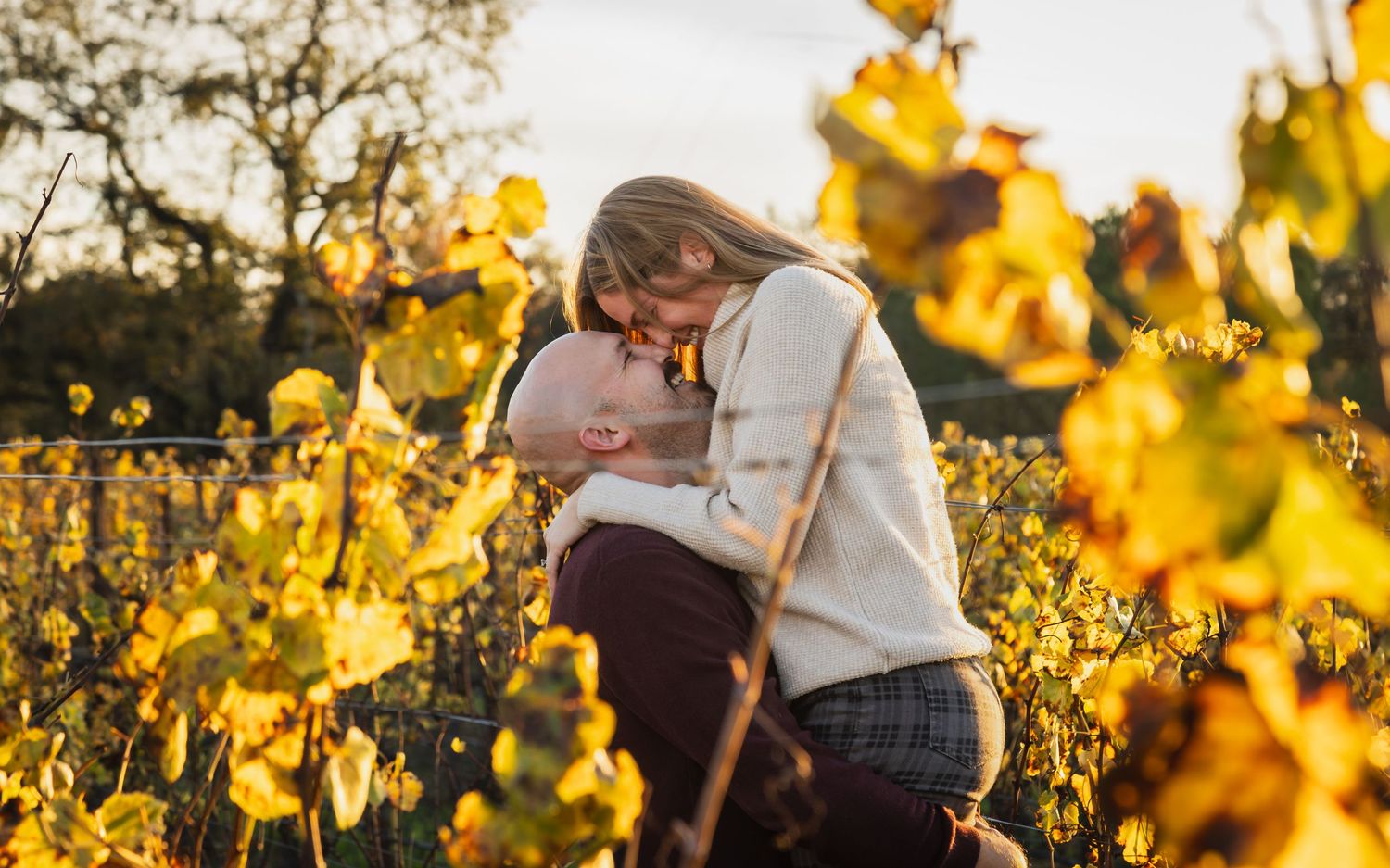A couple shares a loving moment outdoors, surrounded by golden autumn foliage and a serene, rustic backdrop.