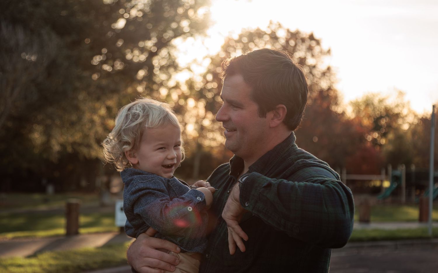 A serene park scene: an adult holds a child, framed by warm sunset light, autumn trees, and a distant playground.