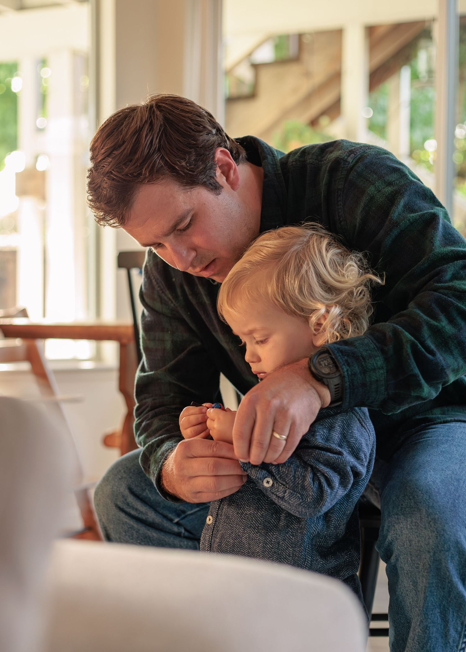 A cozy indoor scene: an adult in a plaid shirt and jeans holds a child's hands, sharing a quiet, intimate moment by a sunlit window.