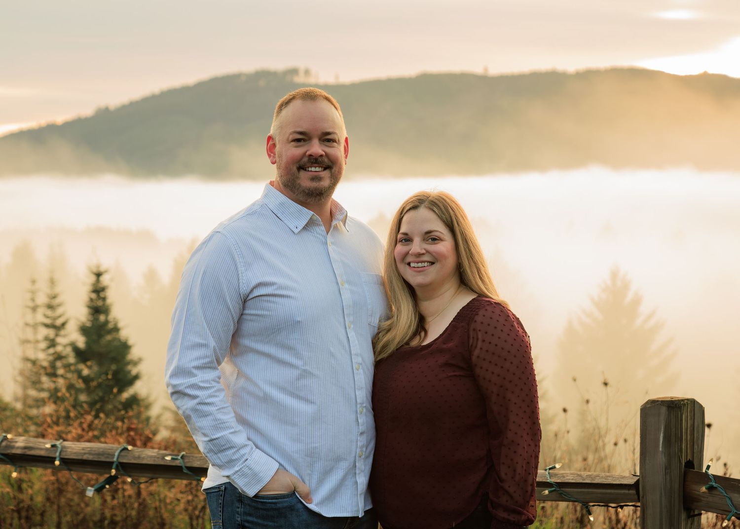 Two people stand close together outdoors, framed by a wooden fence and a misty landscape of hills and trees in soft, golden light.