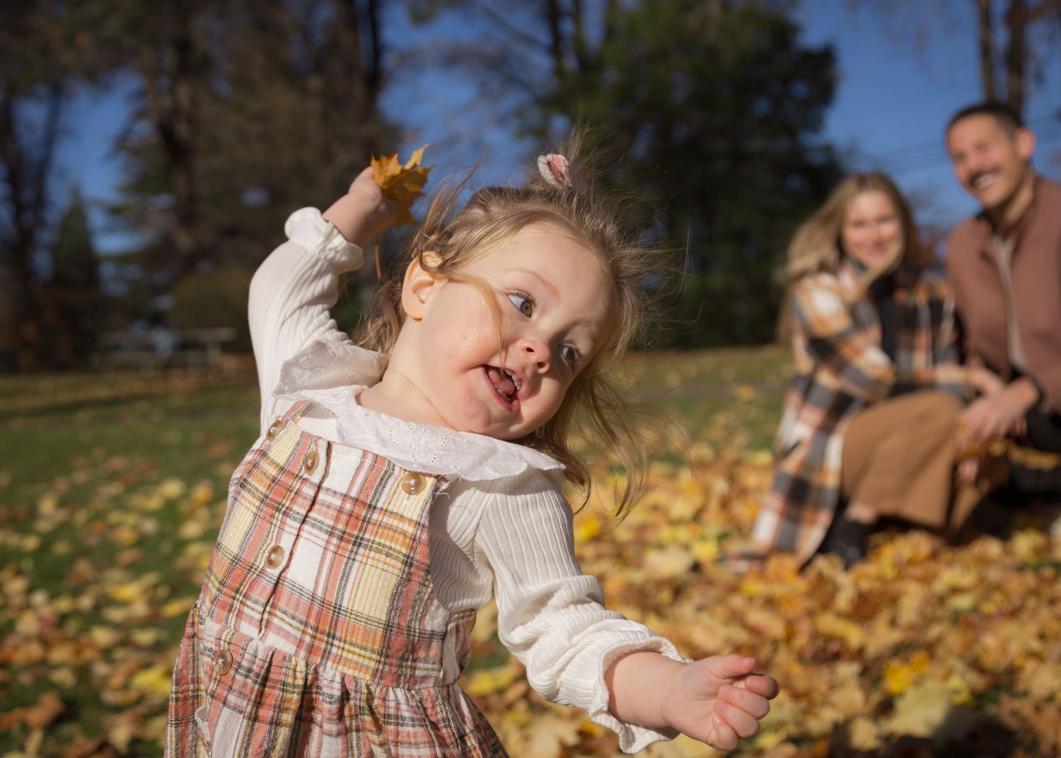 A joyful fall scene: a child plays among leaves, while two adults sit nearby, embracing the warmth of a sunny autumn day.