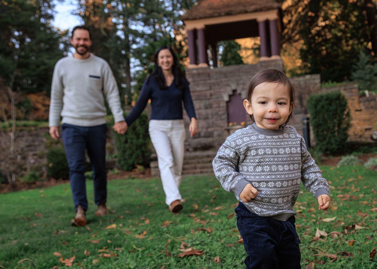 A child in a patterned sweater runs towards the camera in a park, with two adults walking hand-in-hand behind them near a stone structure.