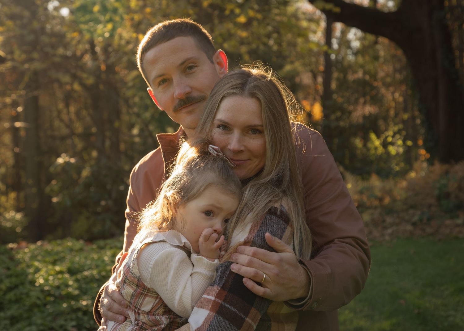 Three people share a warm embrace in a sunlit forest, surrounded by trees and dappled light, evoking closeness and serenity.