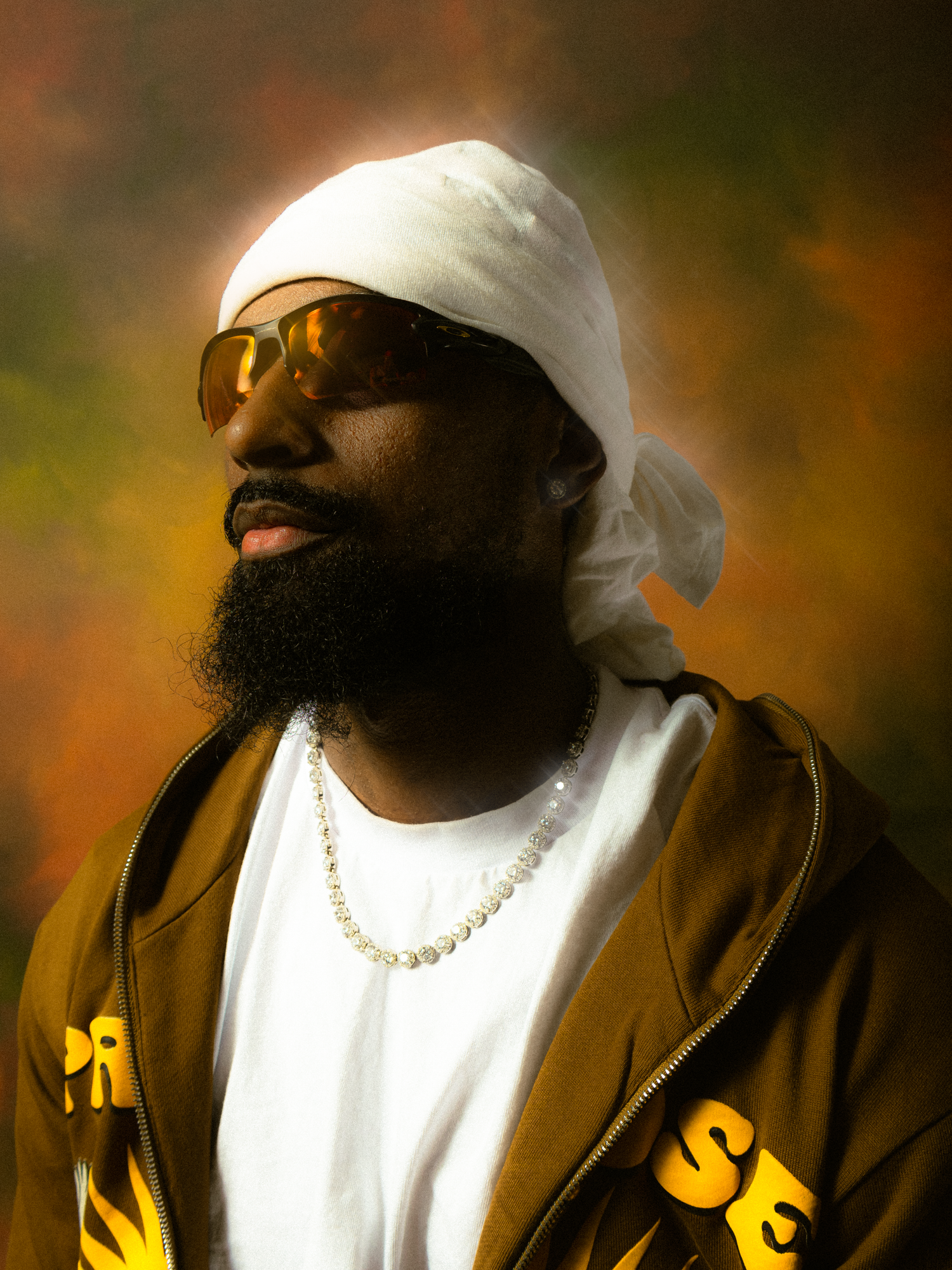 Bearded man wearing white durag, gold sunglasses, pearl necklace, and brown hoodie poses against colorful backdrop.