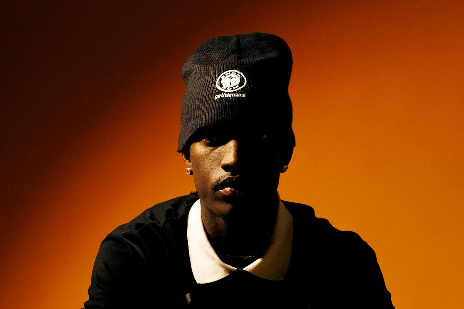 Young man in black beanie hat and polo shirt posed against warm orange gradient background.