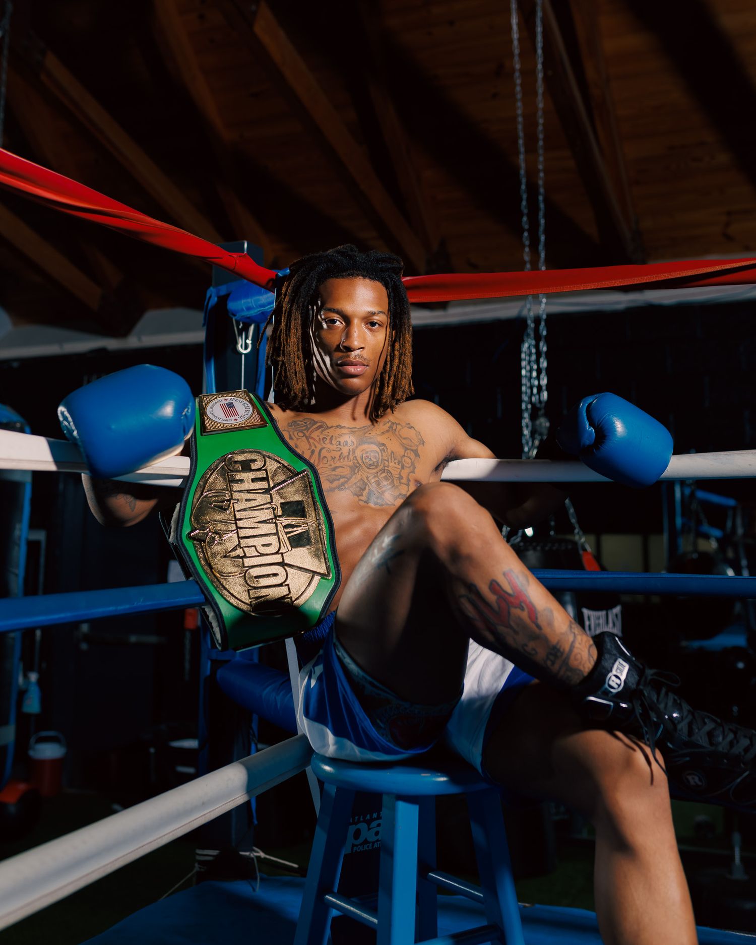 A shirtless boxer with tattoos poses in a ring holding a green champion belt with blue gloves in a gym.