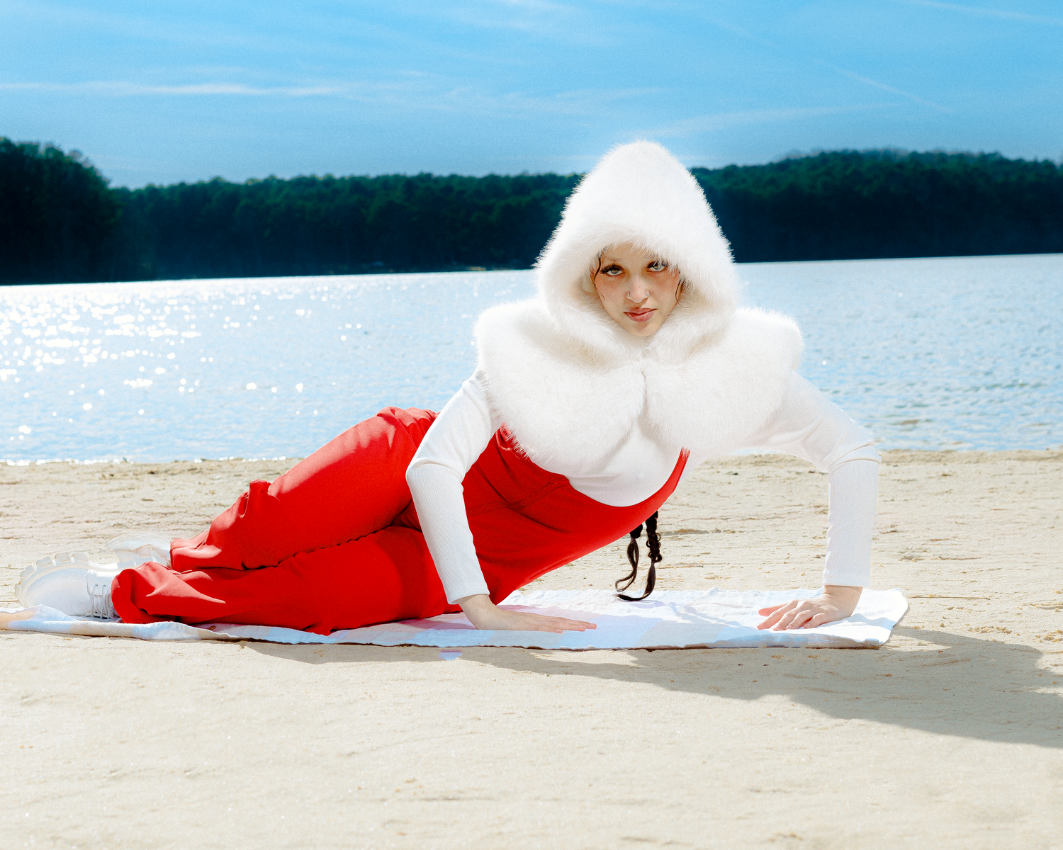 Woman in red skirt and white fur hood poses on sandy beach near sparkling water.