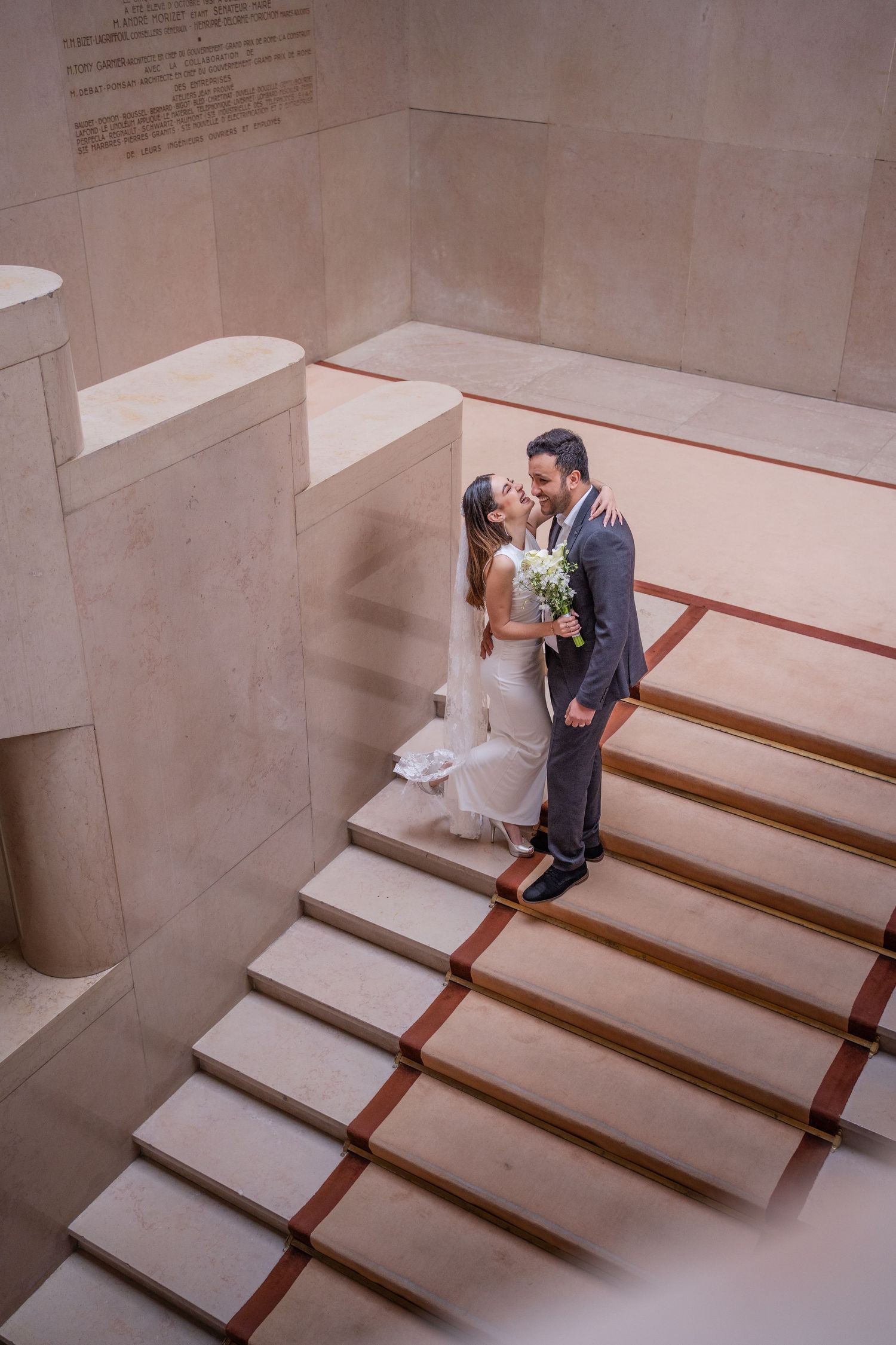 Bride and groom share a kiss on elegant marble stairs, bride holding white bouquet in a stunning wedding venue.