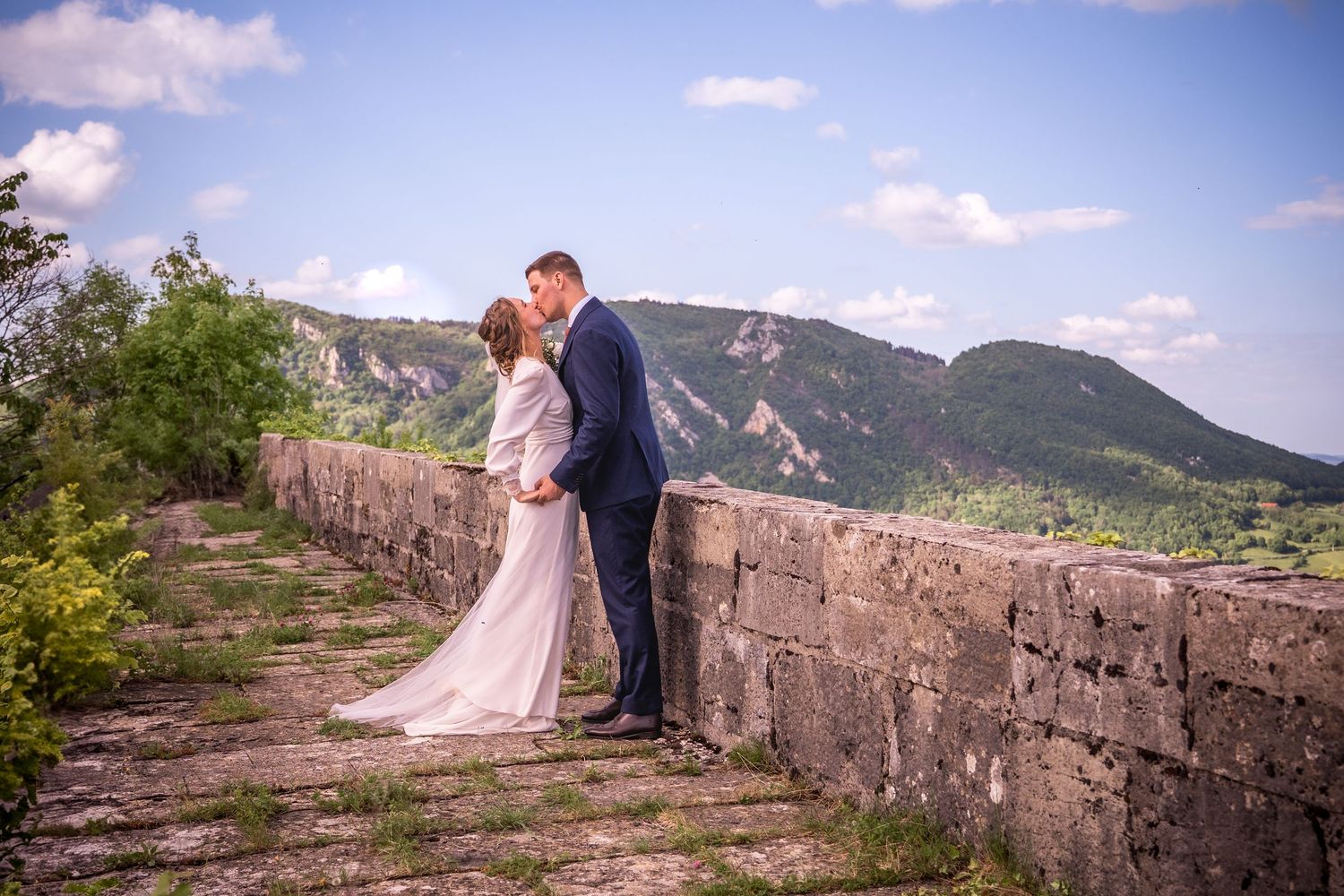 Bride and groom share a romantic kiss on a stone wall with scenic mountain views and blue sky backdrop.