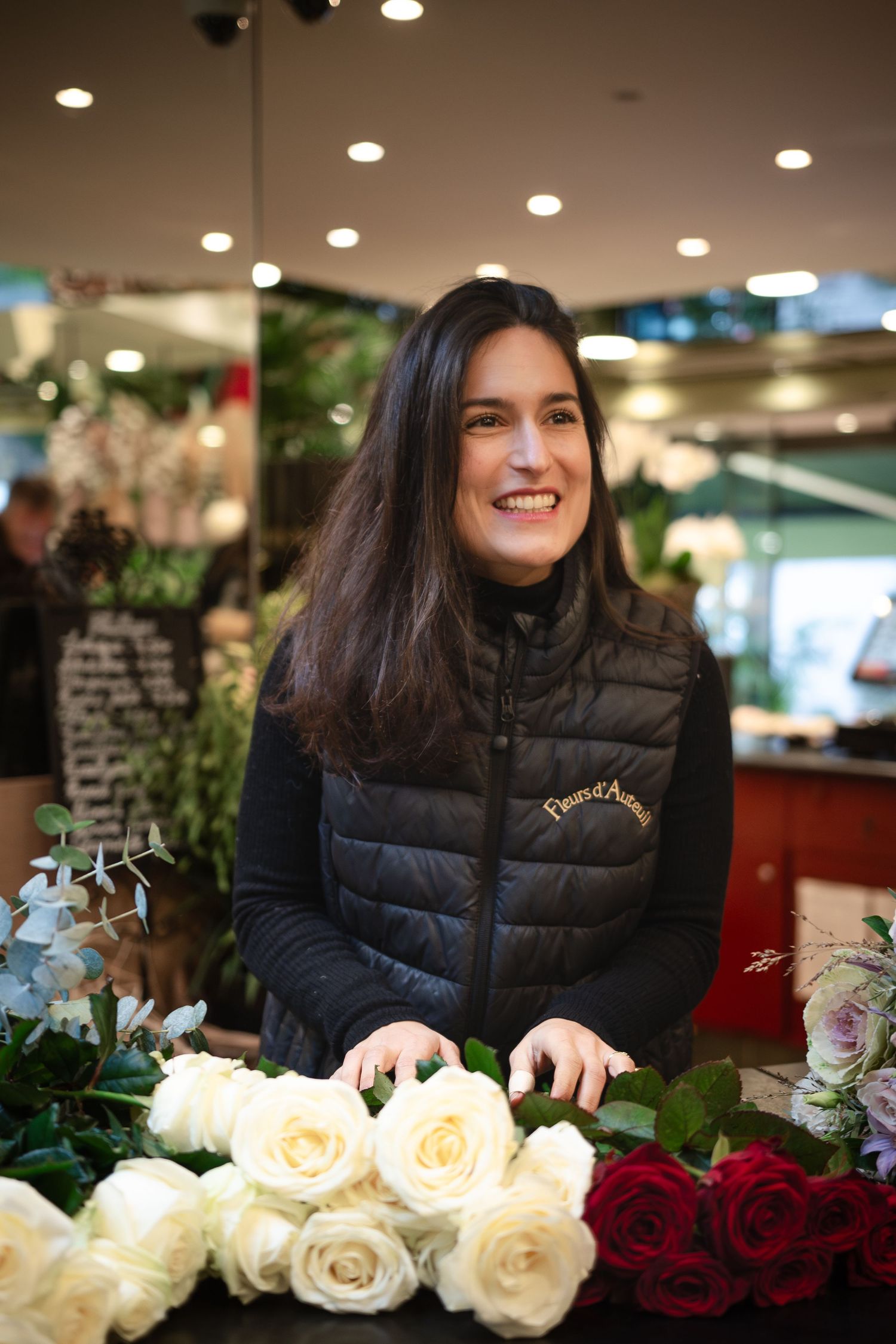 Smiling florist arranging white and red roses at a flower shop counter wearing a black quilted vest.