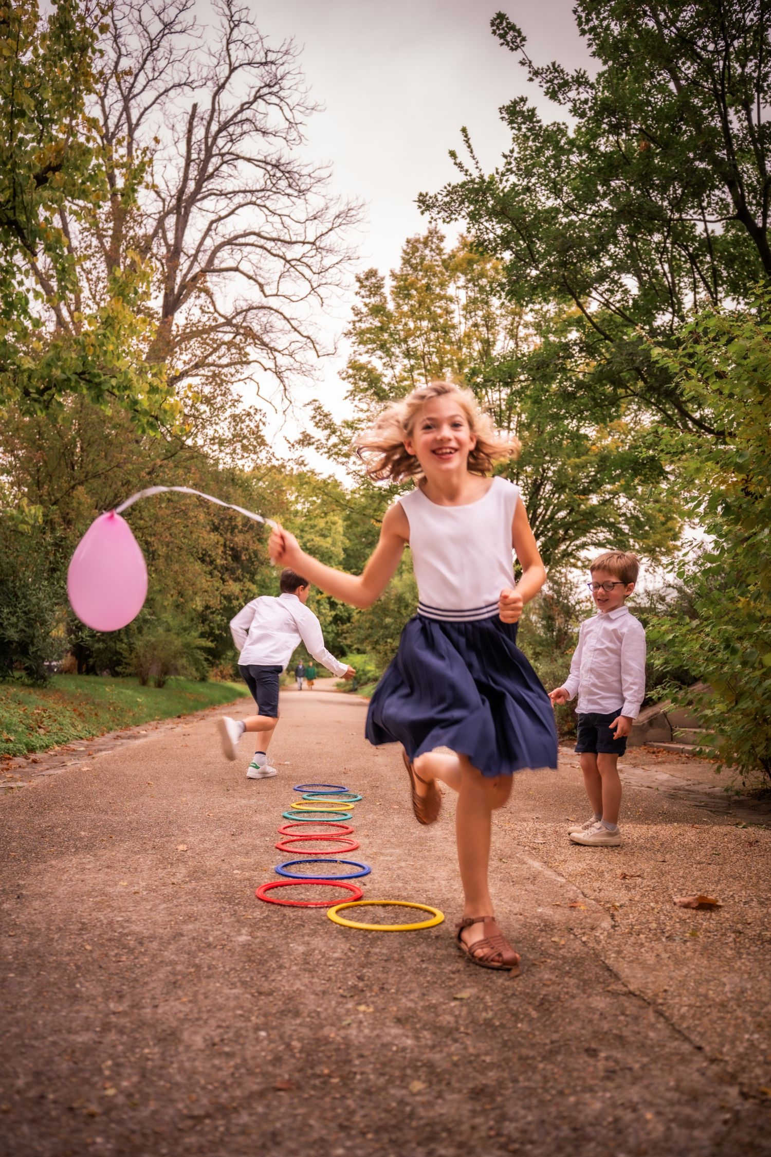 Girl running with pink balloon through colorful hula hoops on park path while children play outdoors.