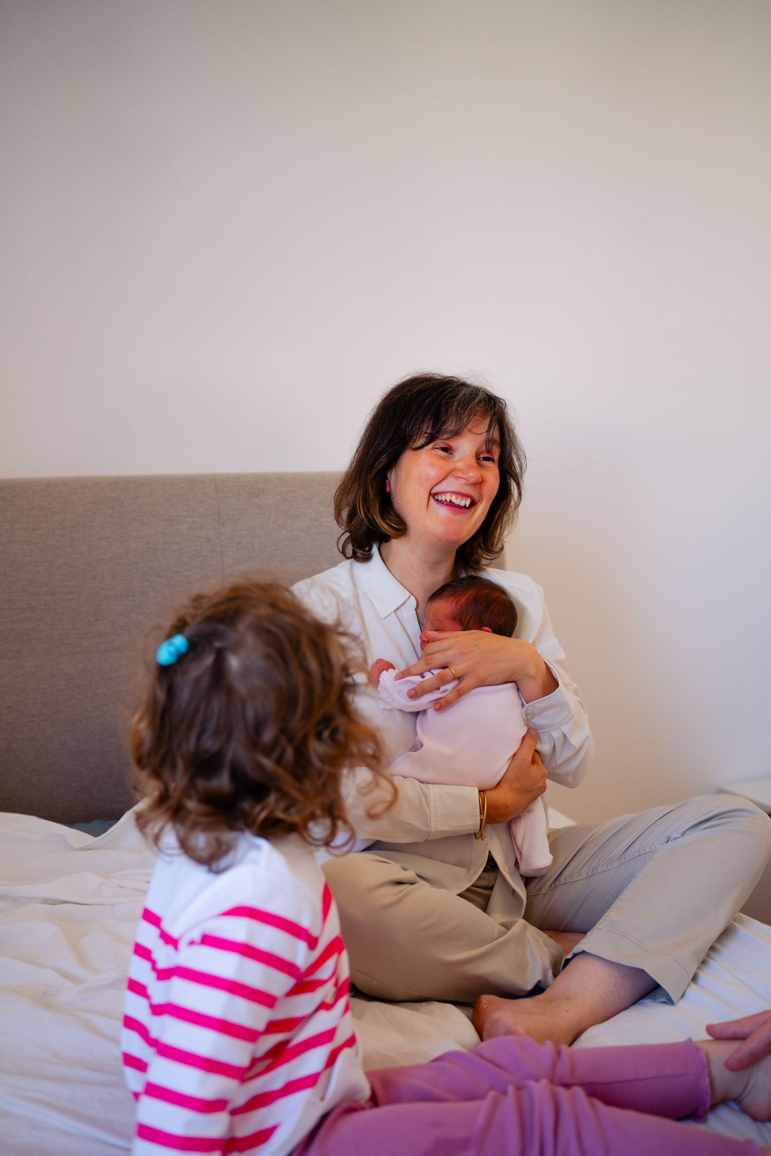 Happy mother sitting on bed holding newborn baby while young toddler daughter watches in cozy bedroom.