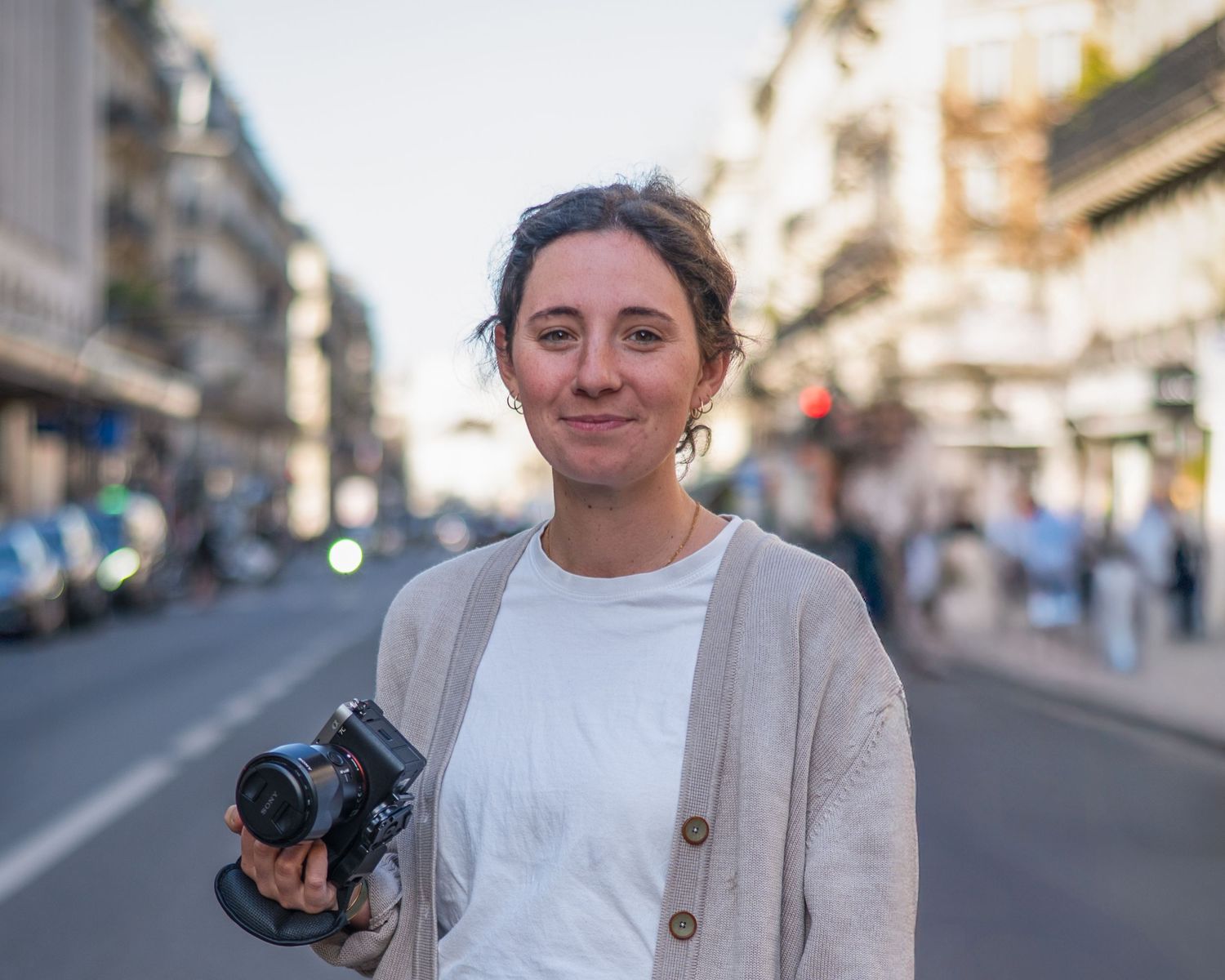 Young woman holding a camera smiles on a busy city street at dusk, with blurred buildings and people in the background.