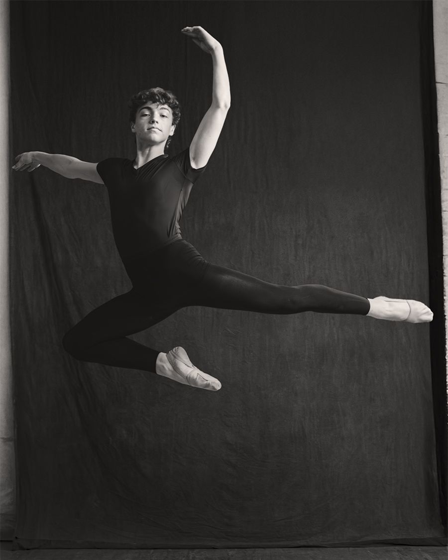 Male ballet dancer leaps gracefully in black attire against dark backdrop in black and white studio photo.