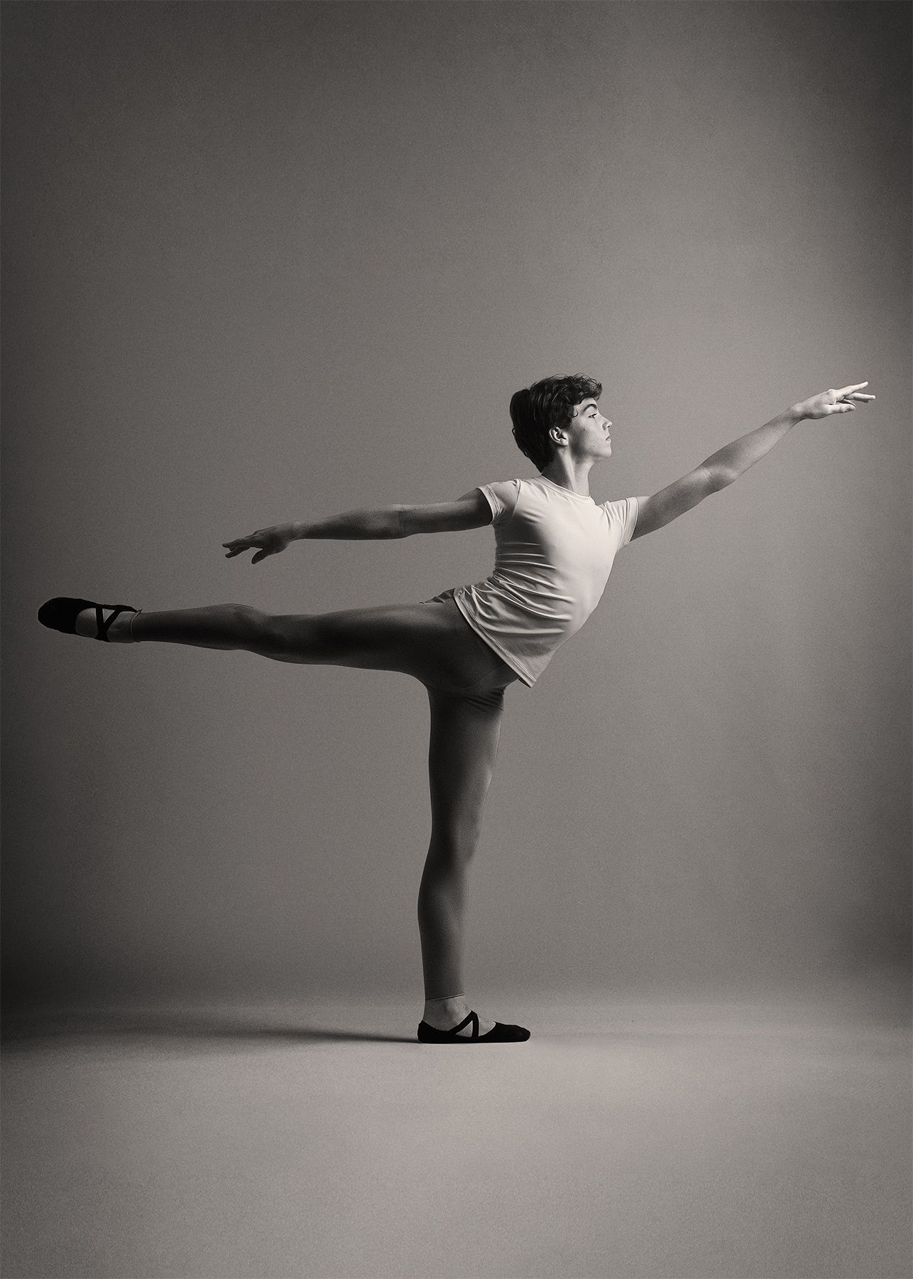 A ballet dancer in a white tank top performs an arabesque pose against a gray background in black and white.