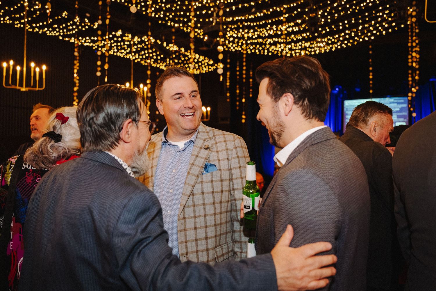 Three men in suits socializing at an elegant indoor event with warm string lights and chandeliers in the background.