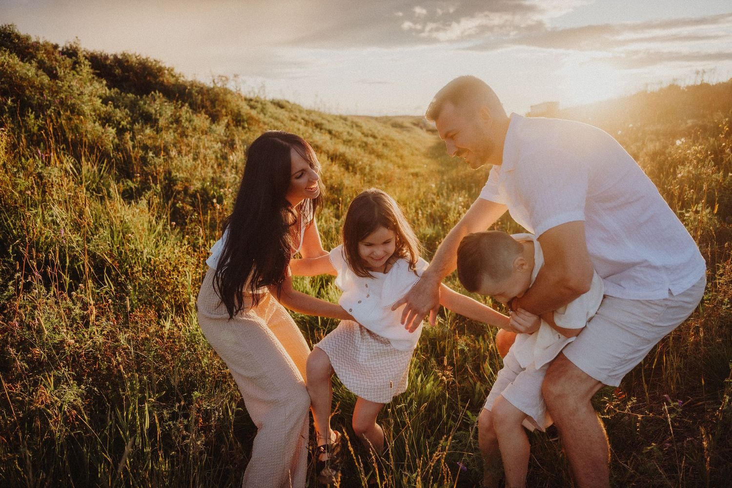 Family in white outfits playing together in a sunlit green meadow at golden hour sunset.