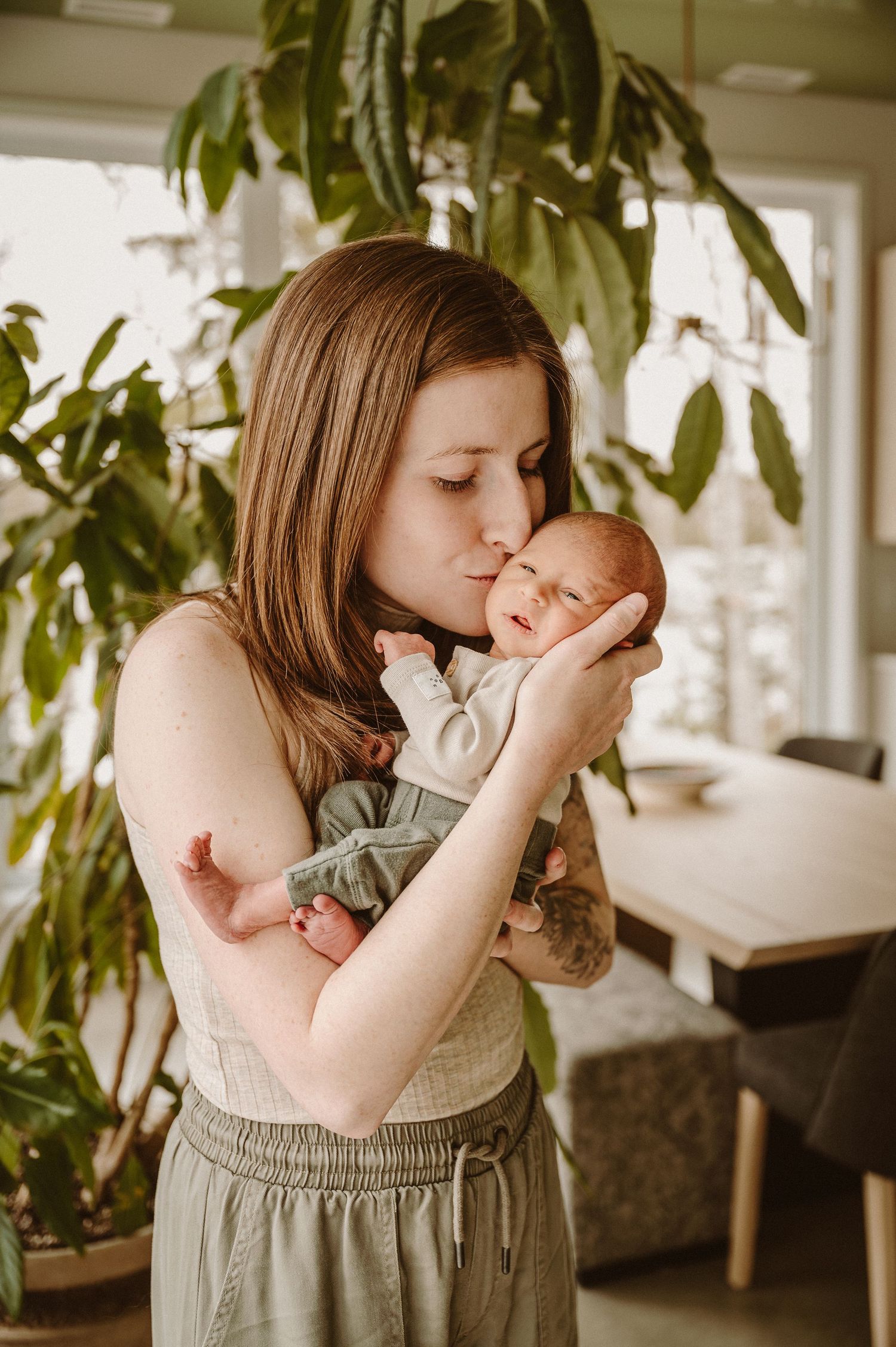 A young mother tenderly holds and kisses her newborn baby indoors, surrounded by lush green plants.