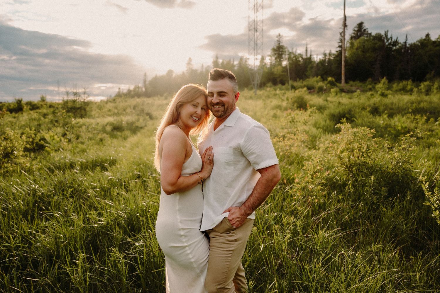 A couple poses romantically in a sunlit green meadow, the woman in white dress leaning into the man in a white shirt.