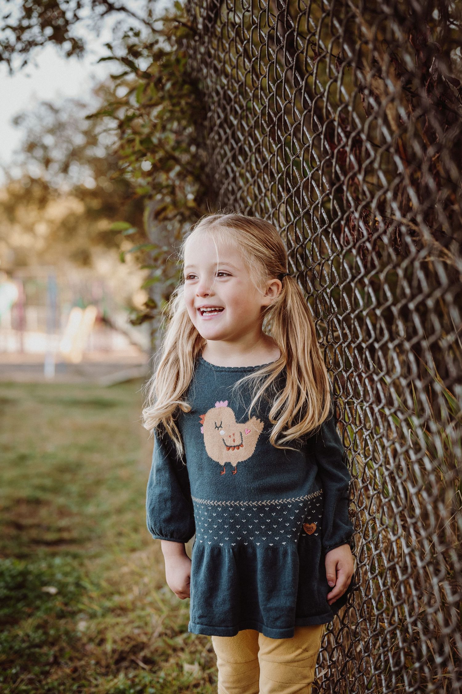 Smiling blonde girl in dark teal top with animal print leans against chain-link fence outdoors in warm autumn light.