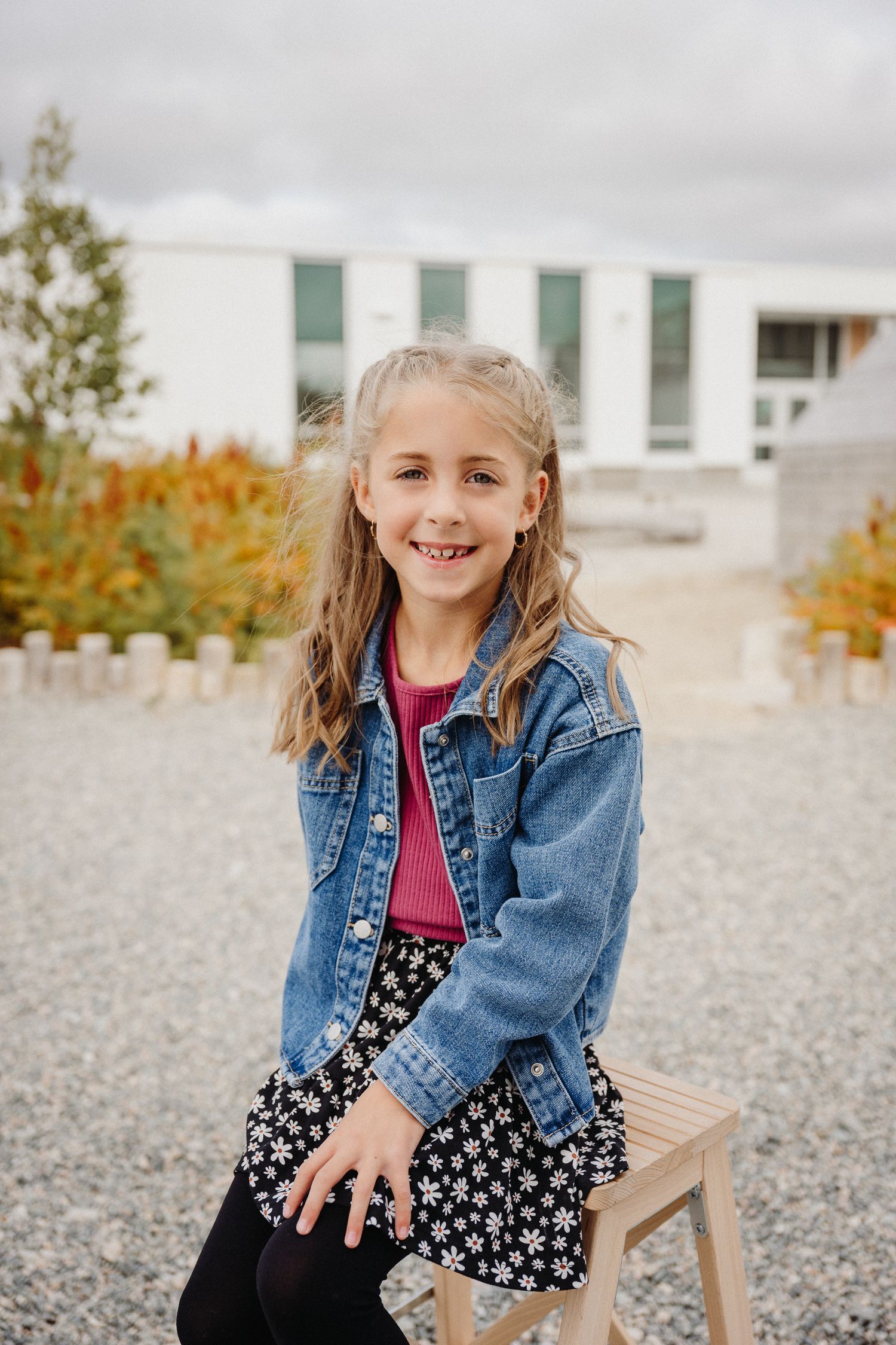A smiling young girl in a denim jacket and floral skirt sits on a wooden stool outdoors near a modern building.