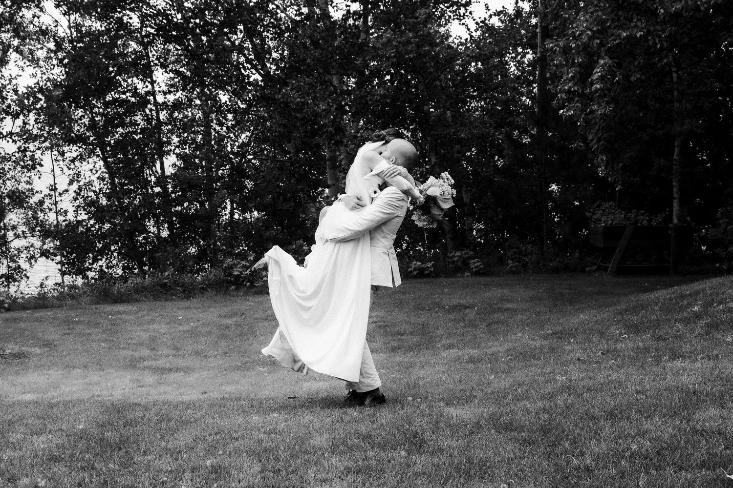 A black and white photograph of a couple dancing romantically on a grassy hillside with trees in the background.