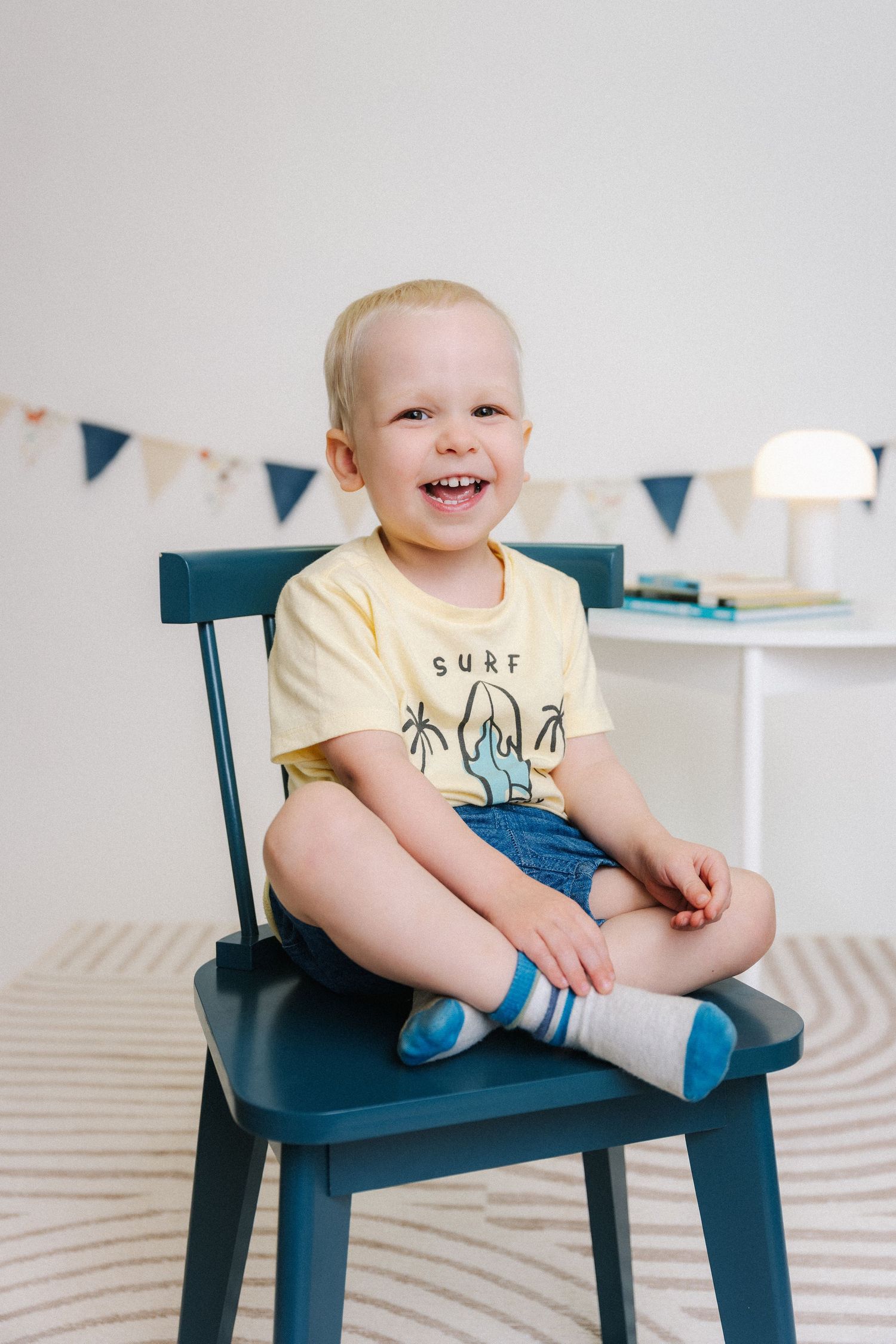 Smiling toddler boy in yellow surf t-shirt and denim shorts sitting cross-legged on a teal wooden chair.