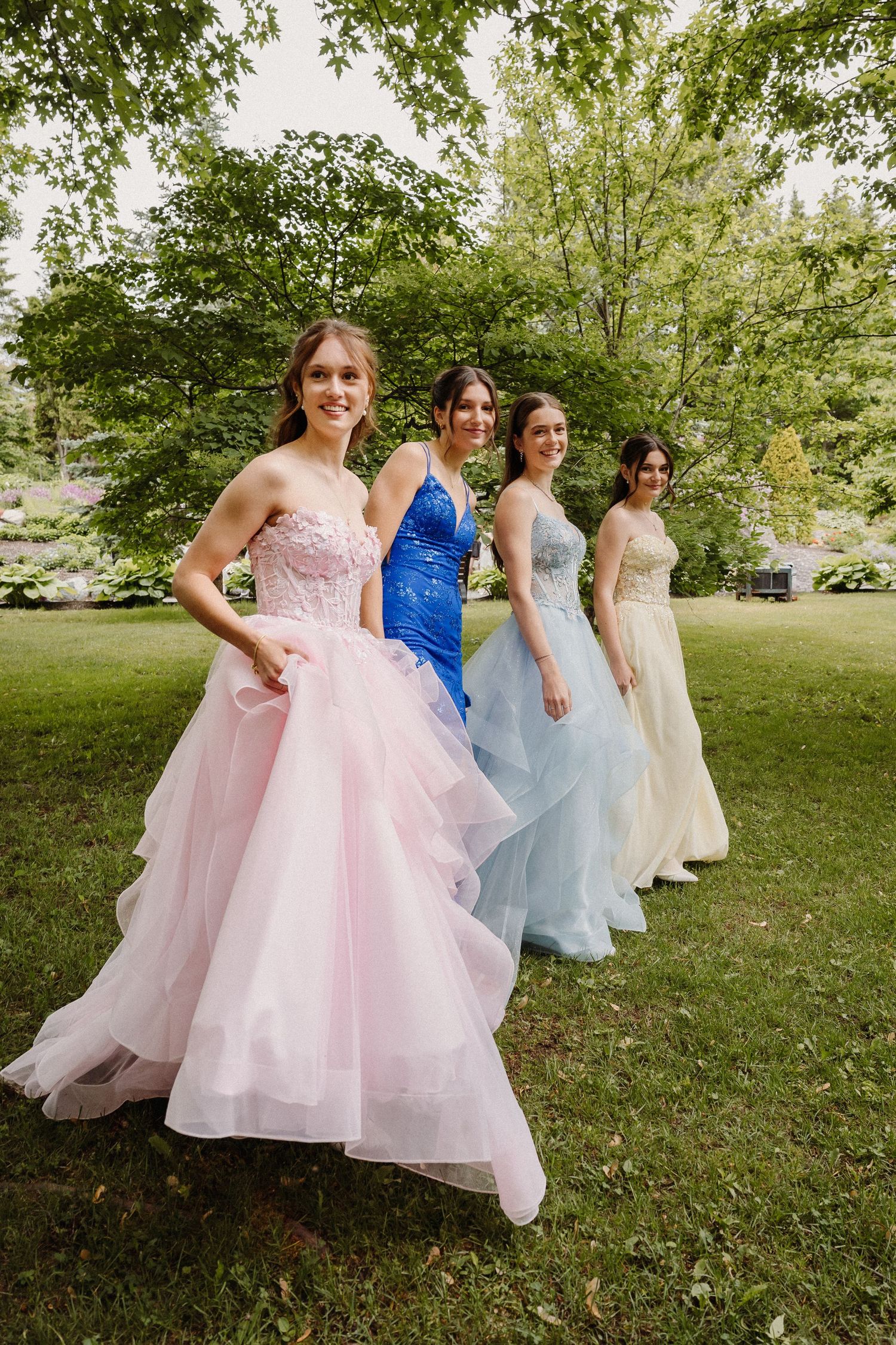 Four young women in elegant prom gowns pose outdoors on green grass surrounded by lush trees.
