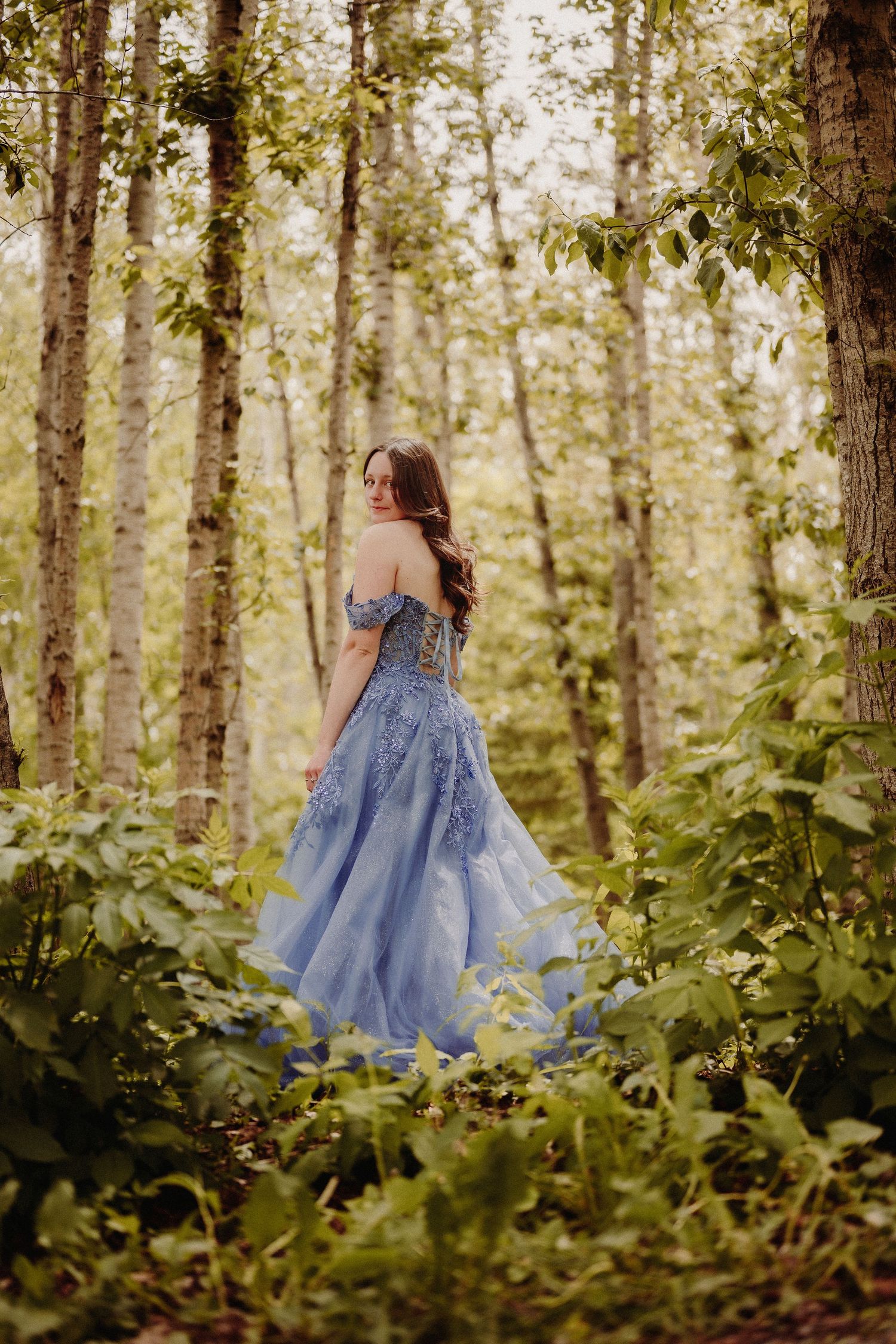 Ethereal blue ball gown flows gracefully through a sunlit forest setting with lush green foliage.