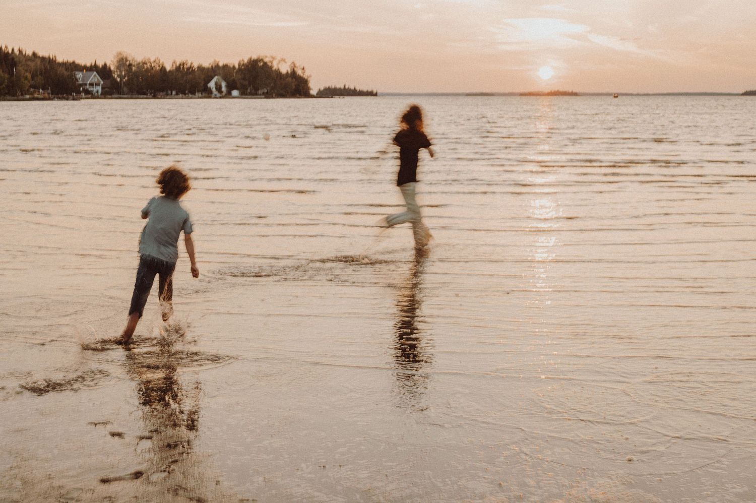 Two silhouettes run and splash along a tranquil beach at sunset with calm water and houses in the distance.