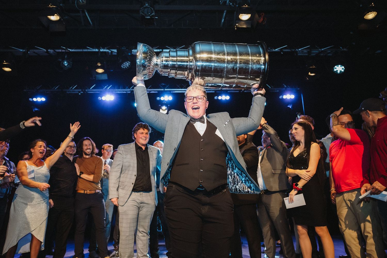 Man triumphantly raises Stanley Cup trophy overhead at celebratory event with cheering crowd behind him.