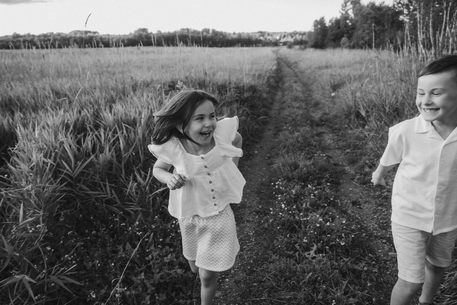 Black and white photo of two happy children running through a field path, girl holding a stuffed animal toy.