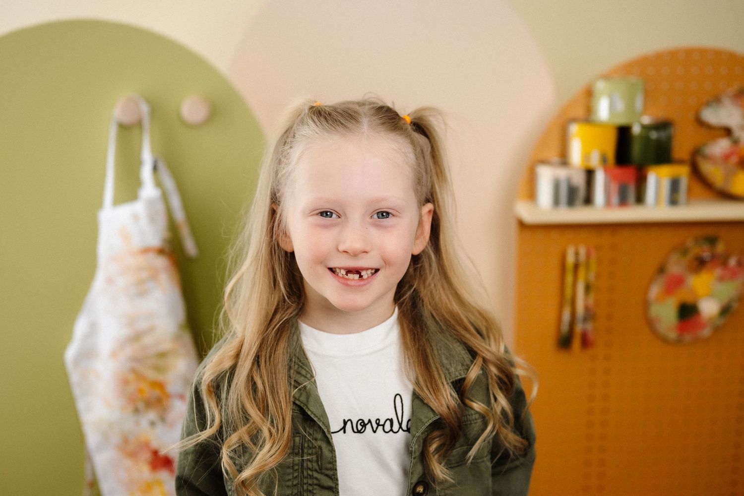 Smiling young girl with pigtails in a green jacket stands in a colorful art room with supplies on shelves.