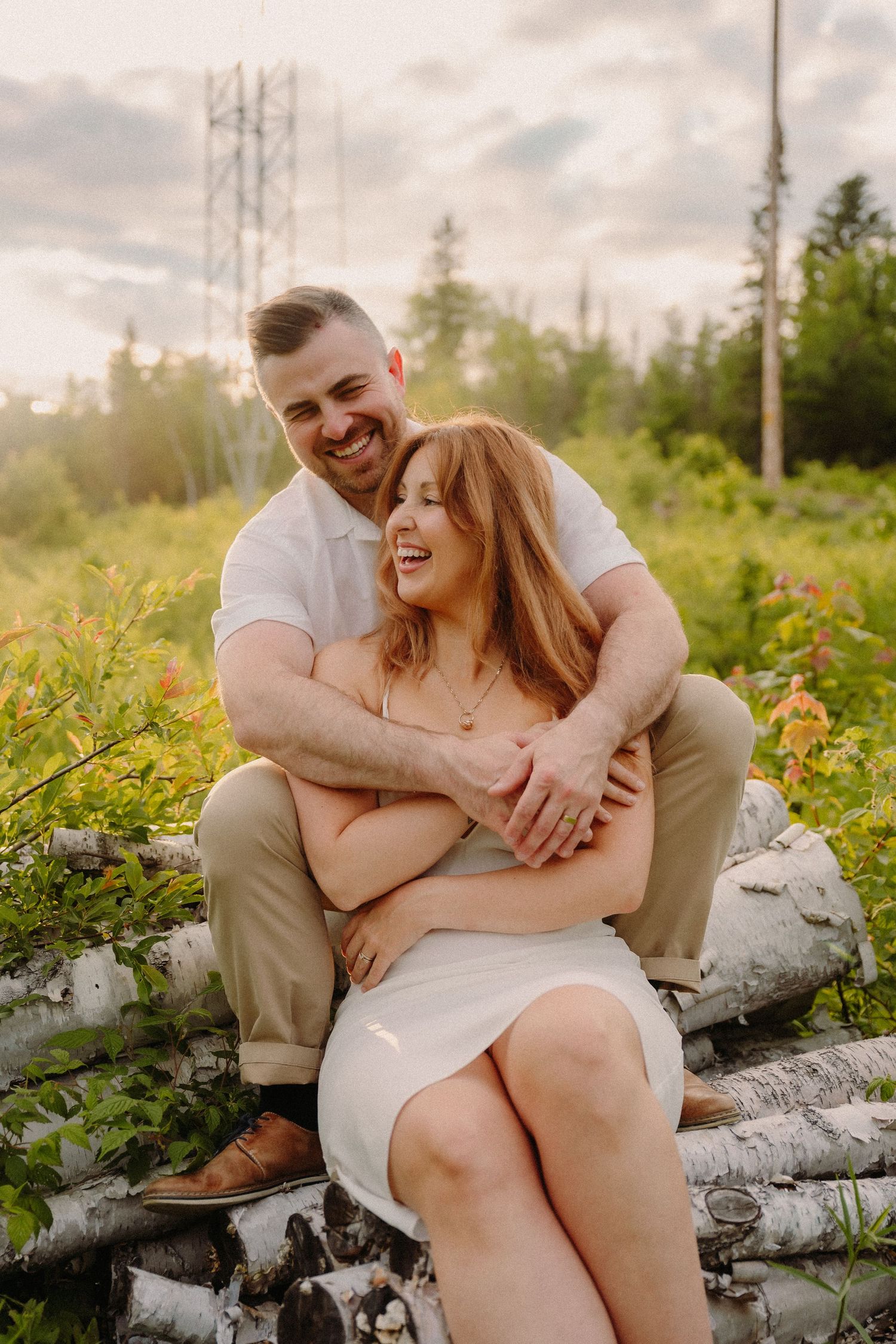 A couple shares a joyful embrace sitting on birch logs in a sunlit meadow, the man hugging the woman from behind.