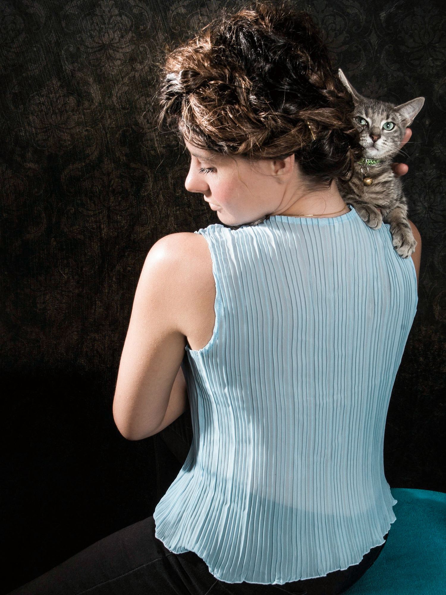 Woman in light blue sleeveless top holding a tabby cat on her shoulder, viewed from behind.