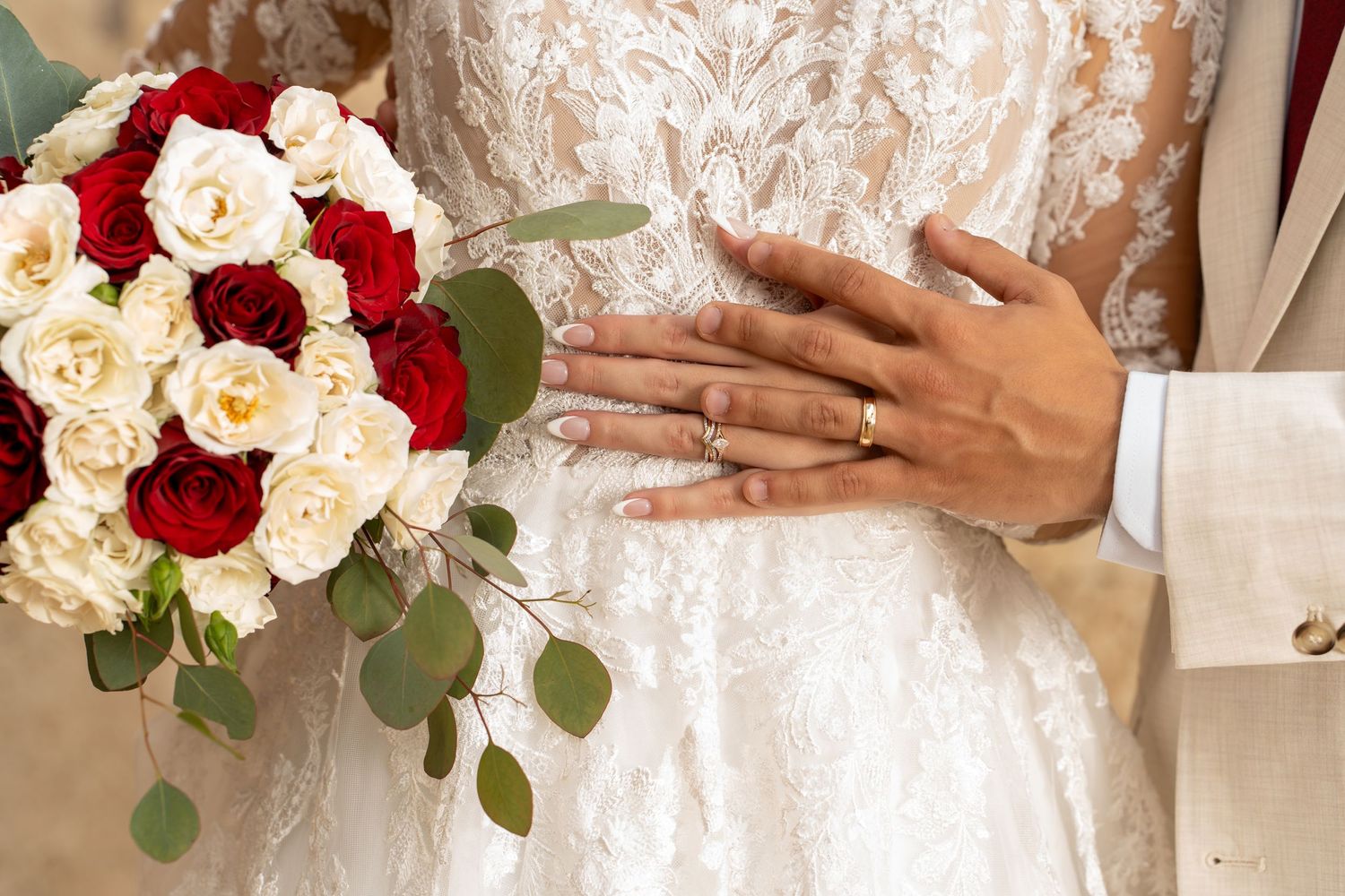 A white and red rose wedding bouquet rests next to hands with wedding rings against a lace dress.