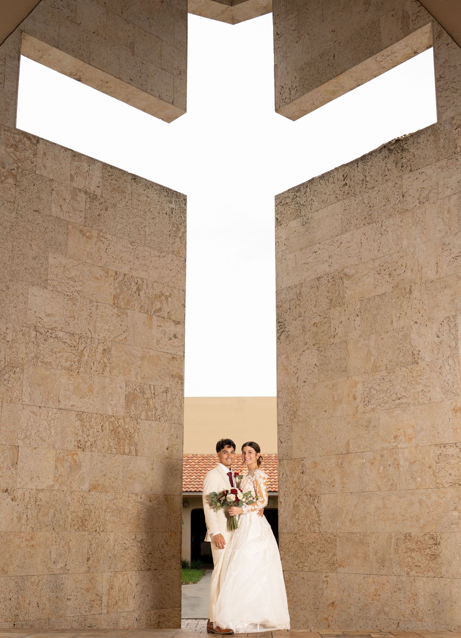 A couple stands under a dramatic stone archway with a cross-shaped opening in white architecture at Catholic church.