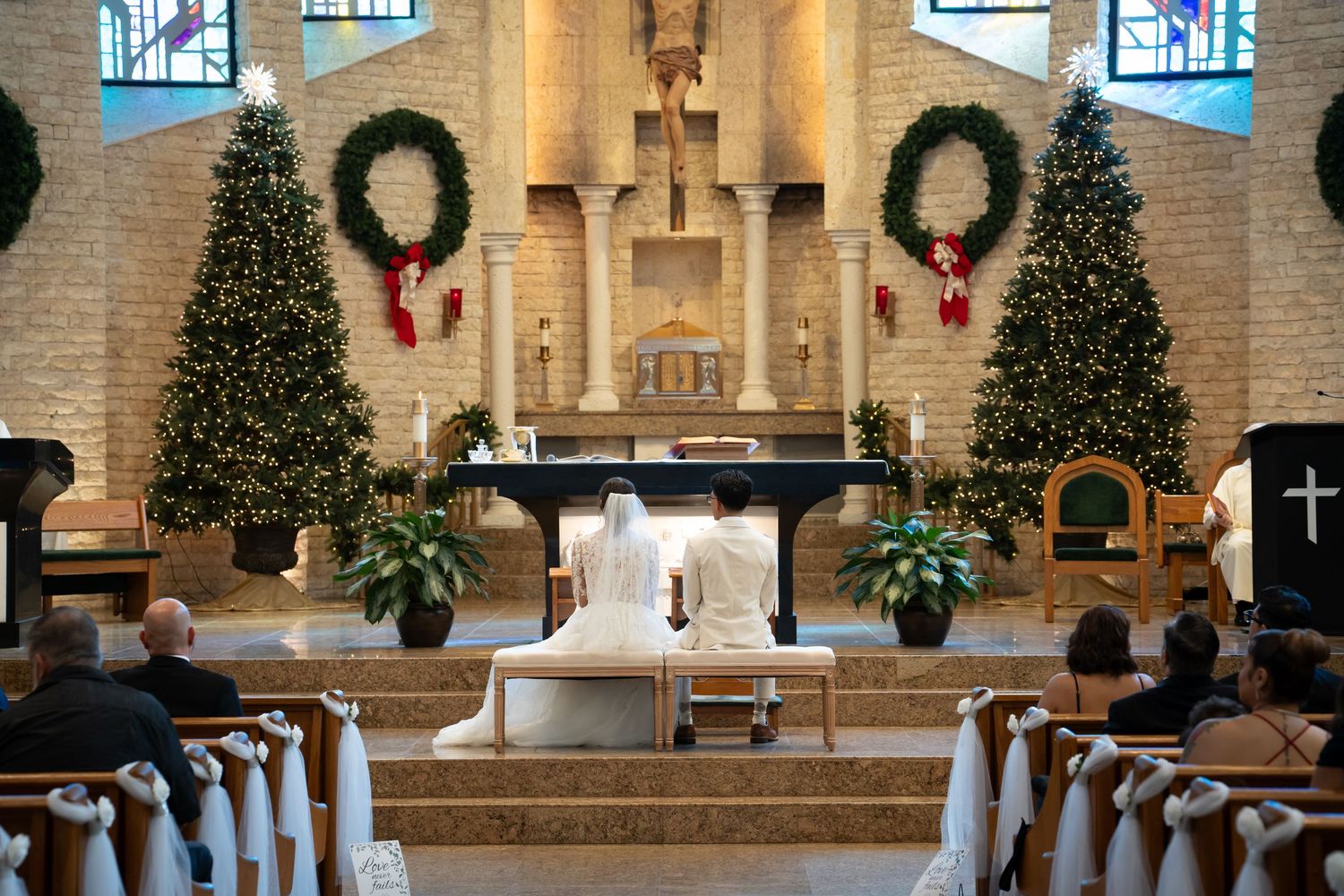 Two decorated Christmas trees flank an altar in a church sanctuary adorned with wreaths and poinsettias for holiday mass.