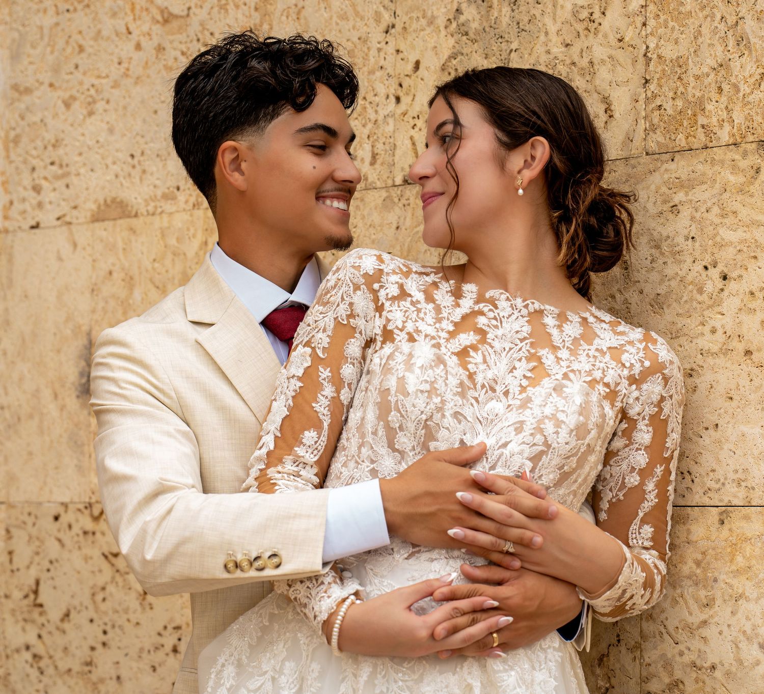 A couple in wedding attire embrace against a textured stone wall, wearing a cream suit and delicate lace dress.