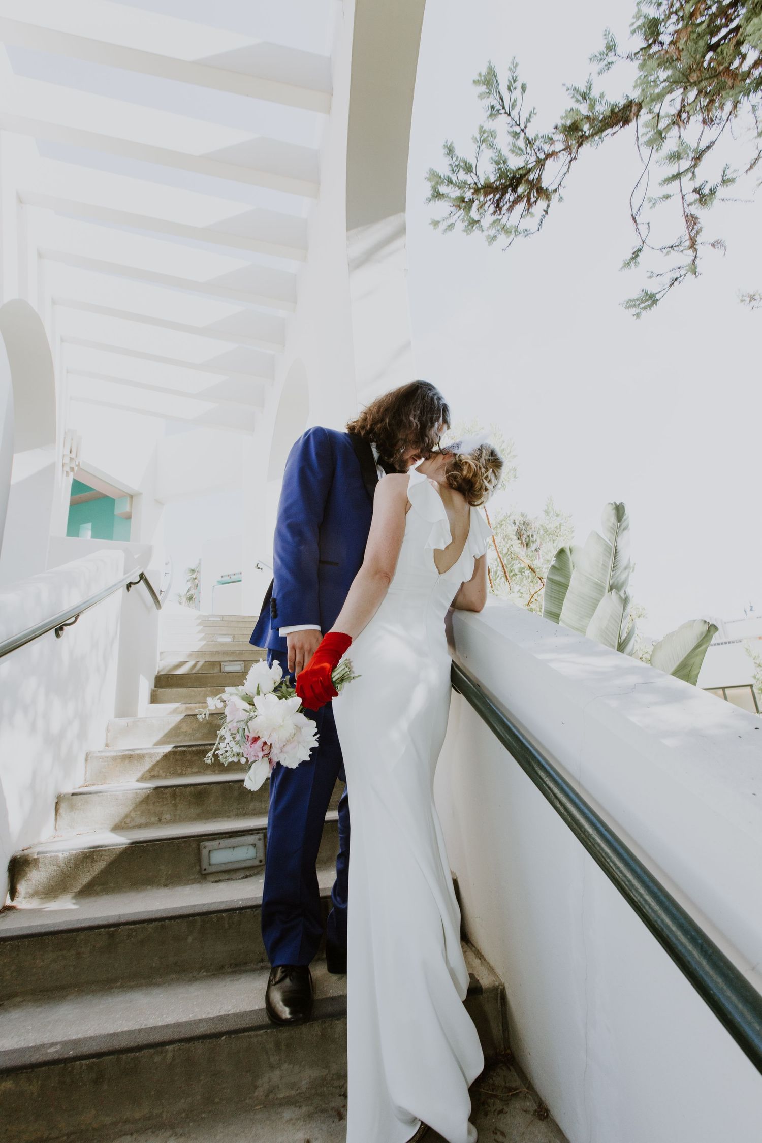 Bride in white gown and red gloves and groom in blue suit share a kiss on sunlit white stairs holding a bridal bouquet.
