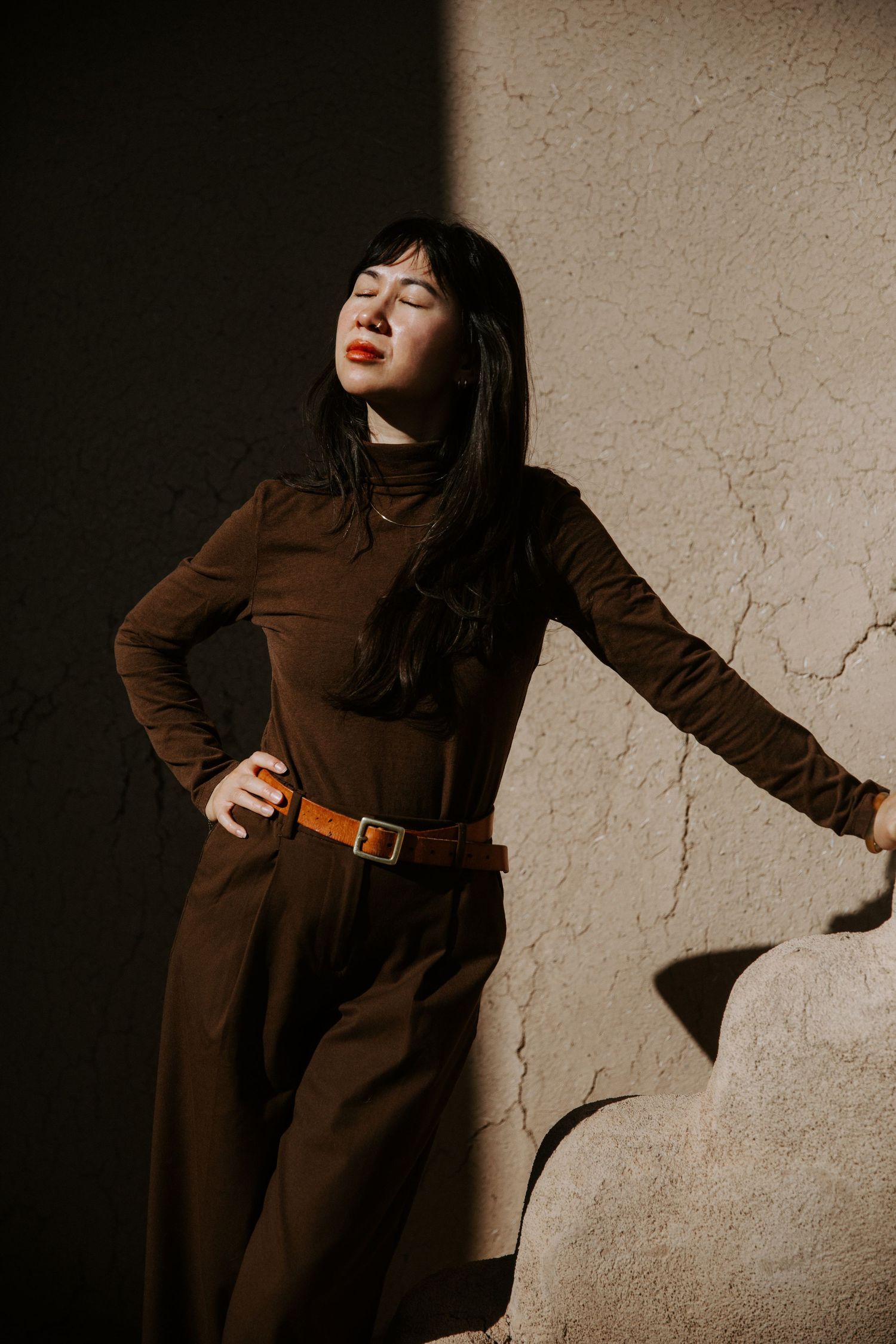 Woman in brown turtleneck jumpsuit with tan belt poses against textured wall in dramatic sunlight.